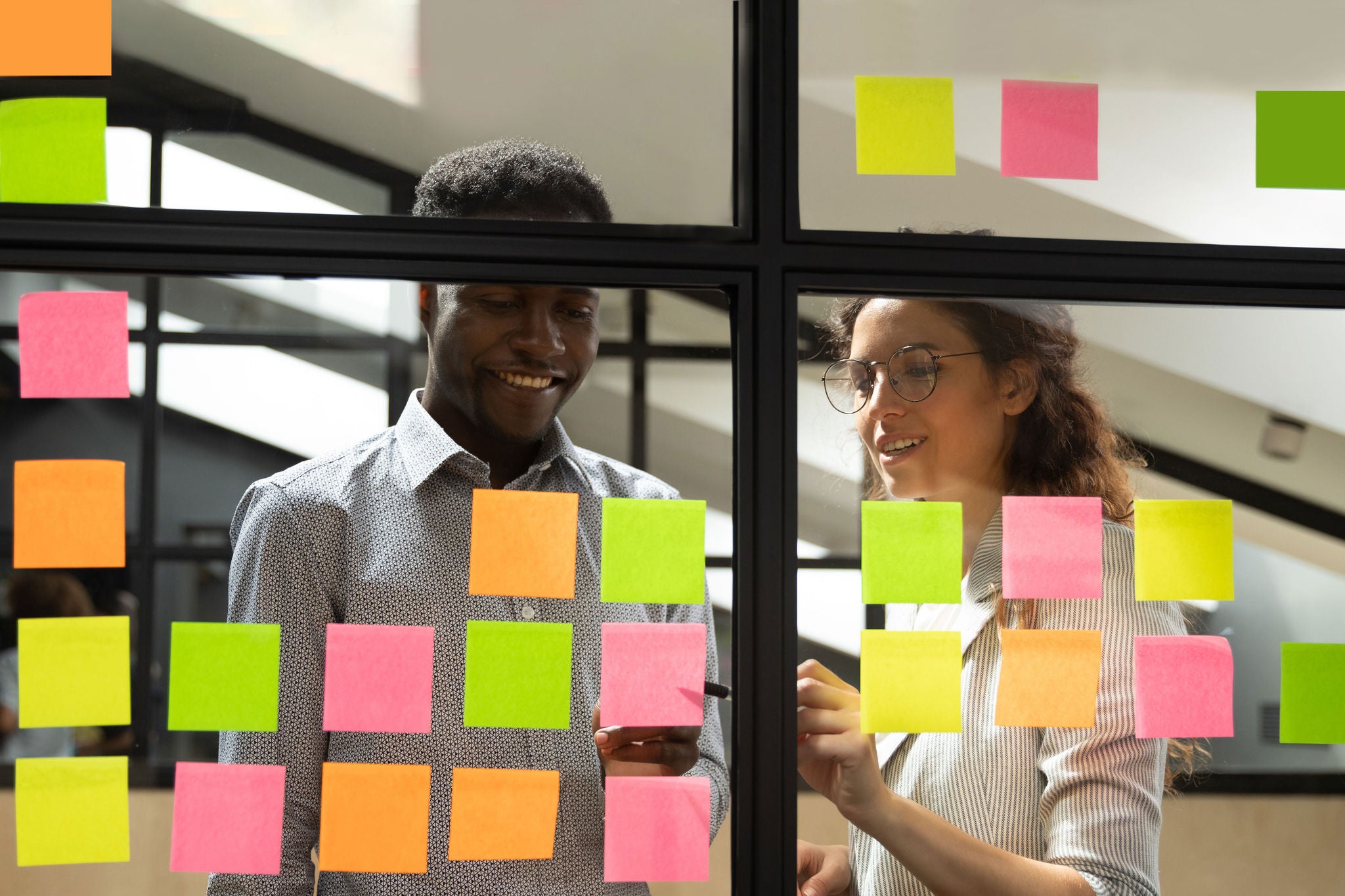 Multiracial colleagues developing business ideas use post-it notes view through glass, mates read written tasks shown on stickers attached on wall, effective way do work don't forget anything concept