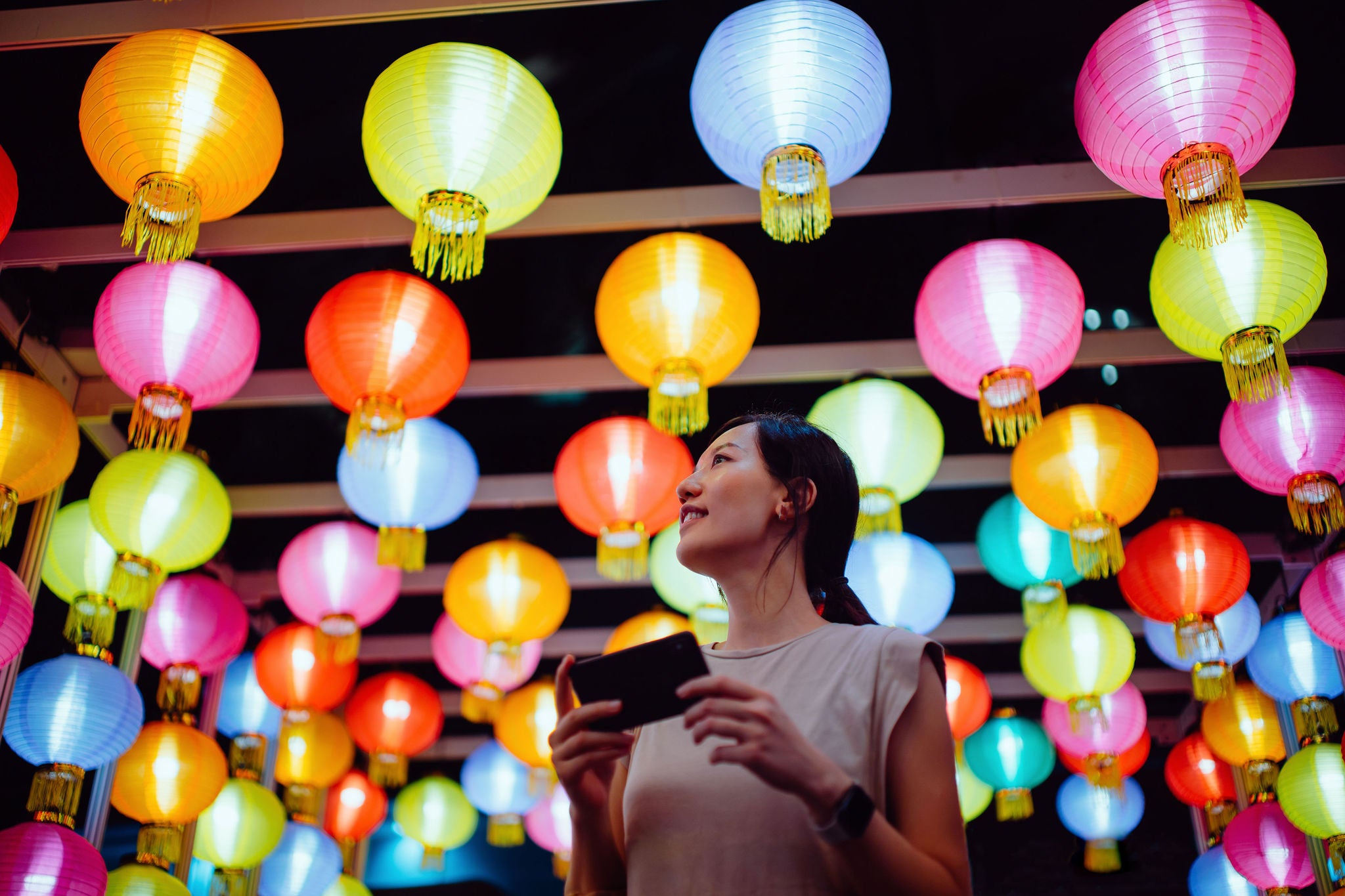Smiling young Asian woman taking photos of illuminated and colourful traditional Chinese lanterns with smartphone hanging on city street at night. Traditional Chinese culture, festival and celebration event theme