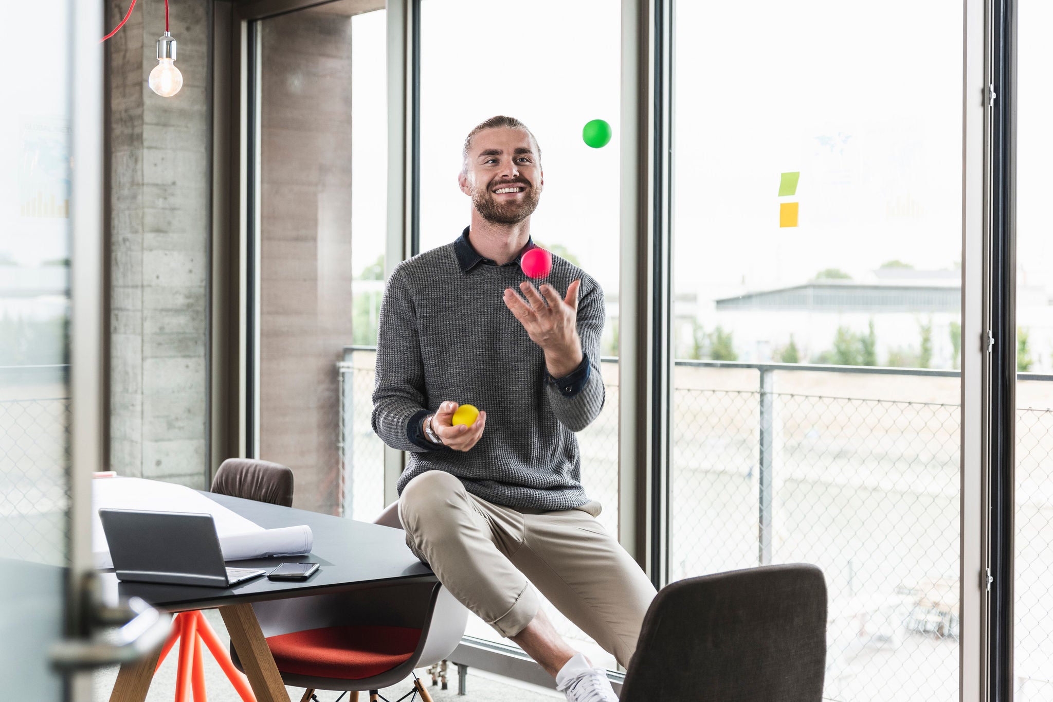 Smiling young businessman sitting at the window juggling with balls