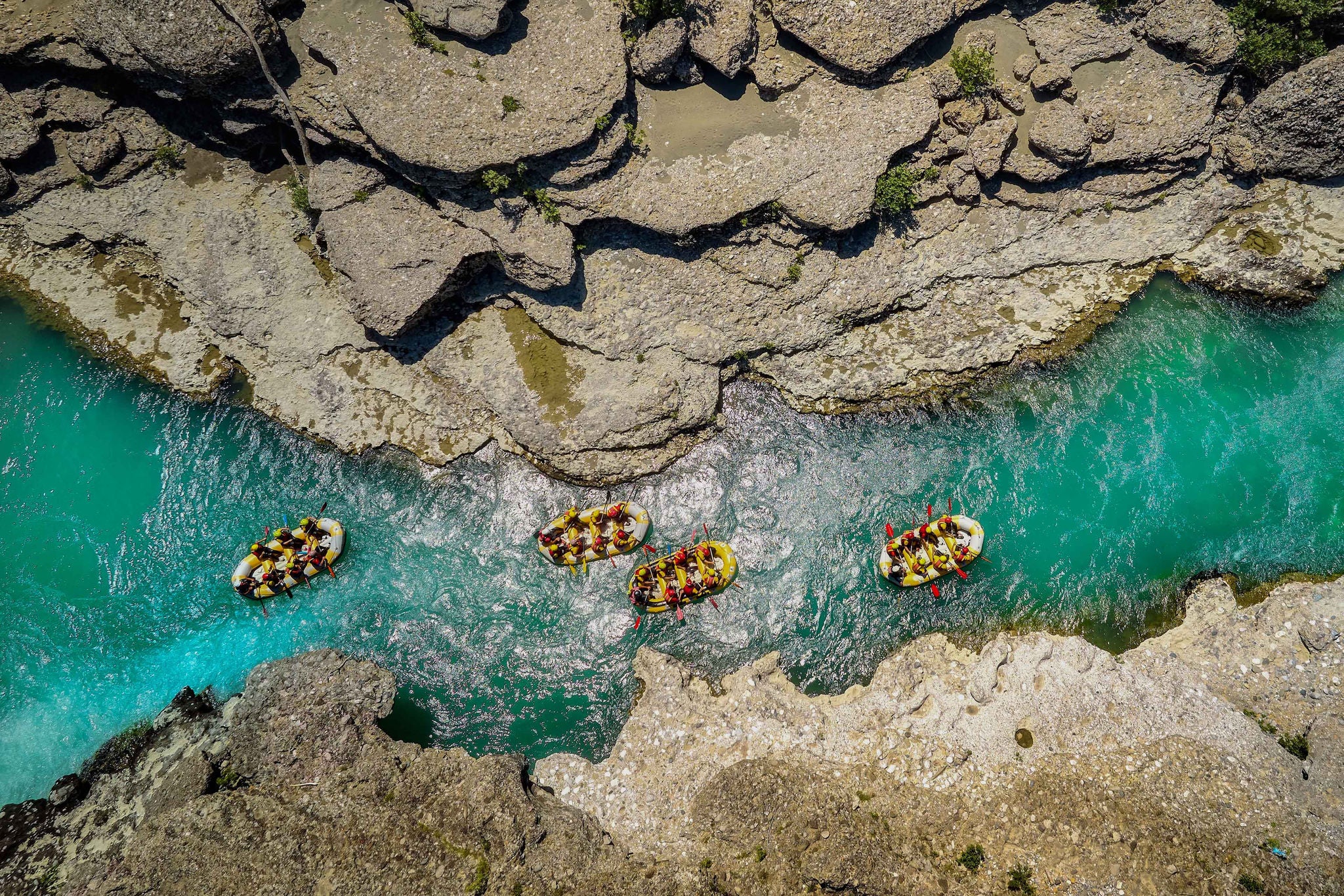 Four yellow rafts floating among the rocks on the crystal clear, blue-green water. Aerial view of the rafters floating on Vjosa river. White water rafting.  Adventure and sport