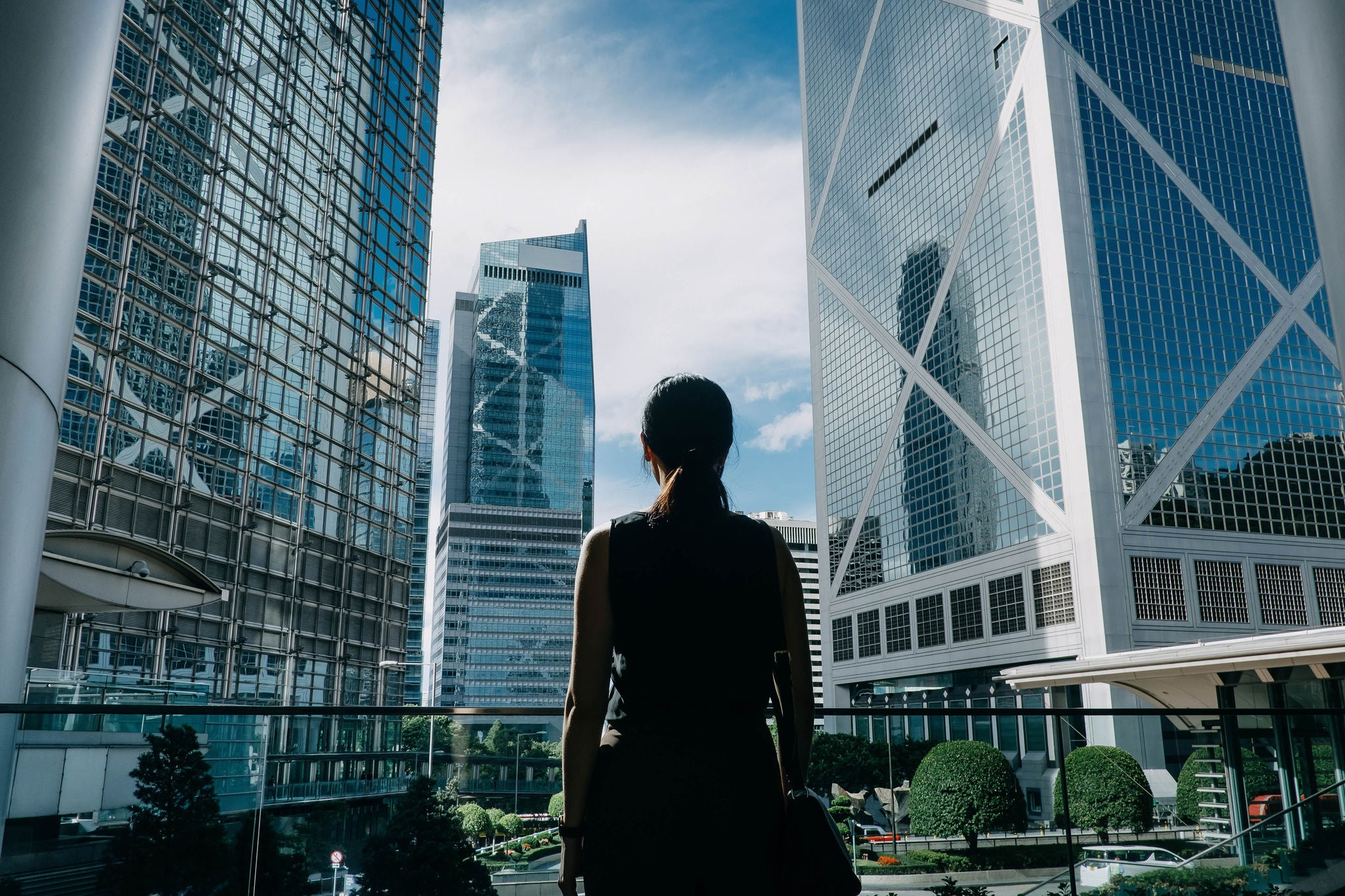 Rear view of professional young businesswoman standing against contemporary financial skyscrapers in downtown financial district and looking up into sky with positive emotion