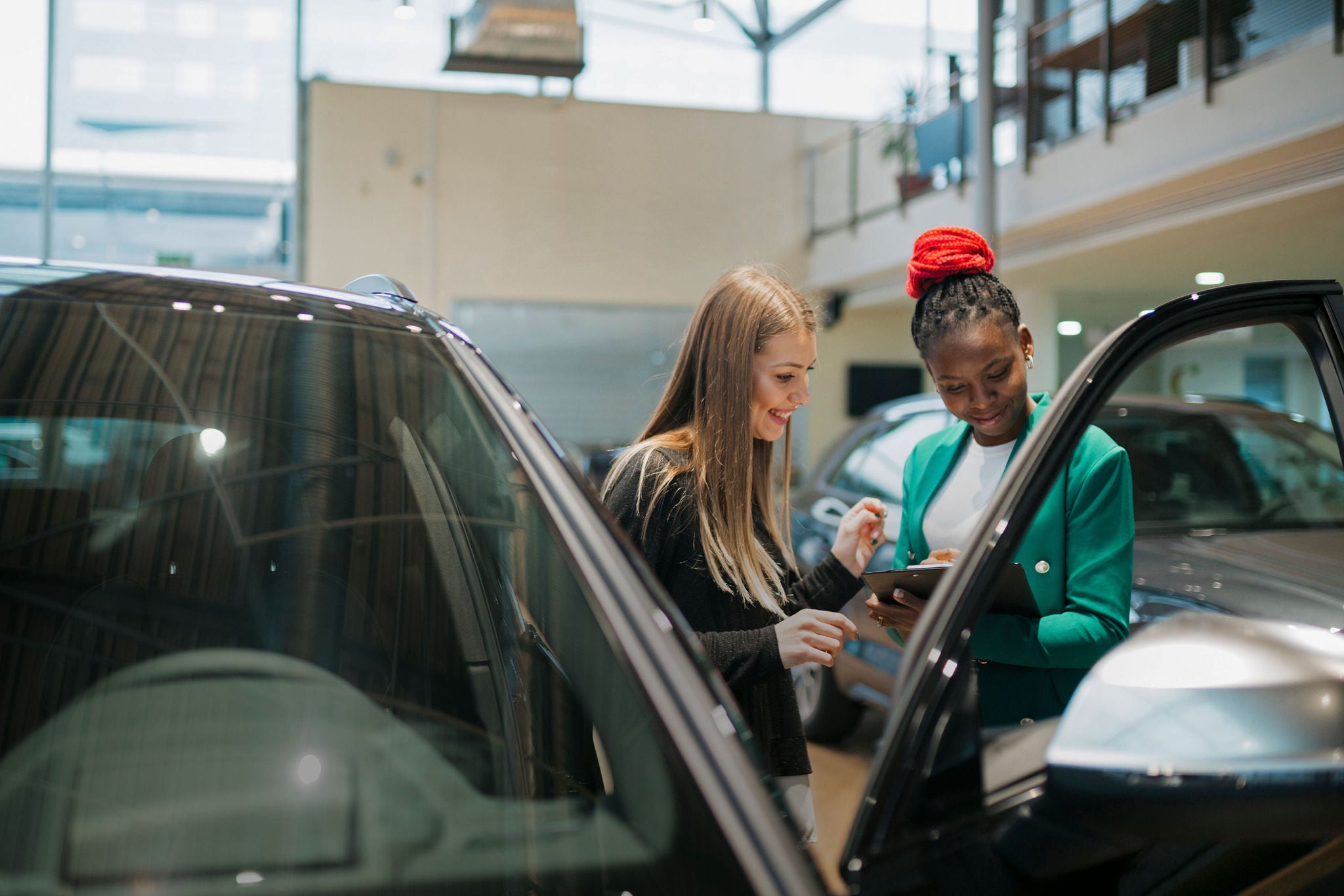 Young car saleswoman talking to a female customer at the showroom. Sales manager sharing the car details looking at a clipboard with woman customer at auto dealership.