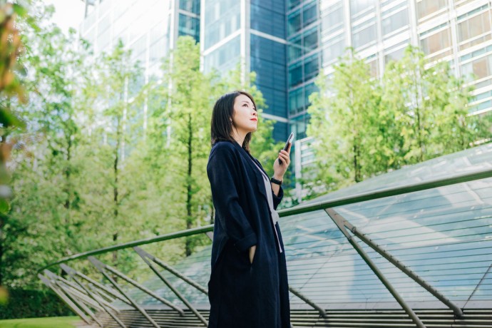 A young asian woman holding a smart phone standing against corporate buildings