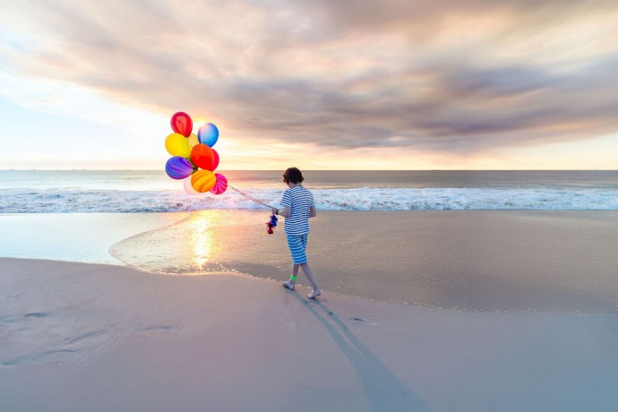Boy with a bunch of colorful balloons, walking along the water's edge at the beach as the sun sets