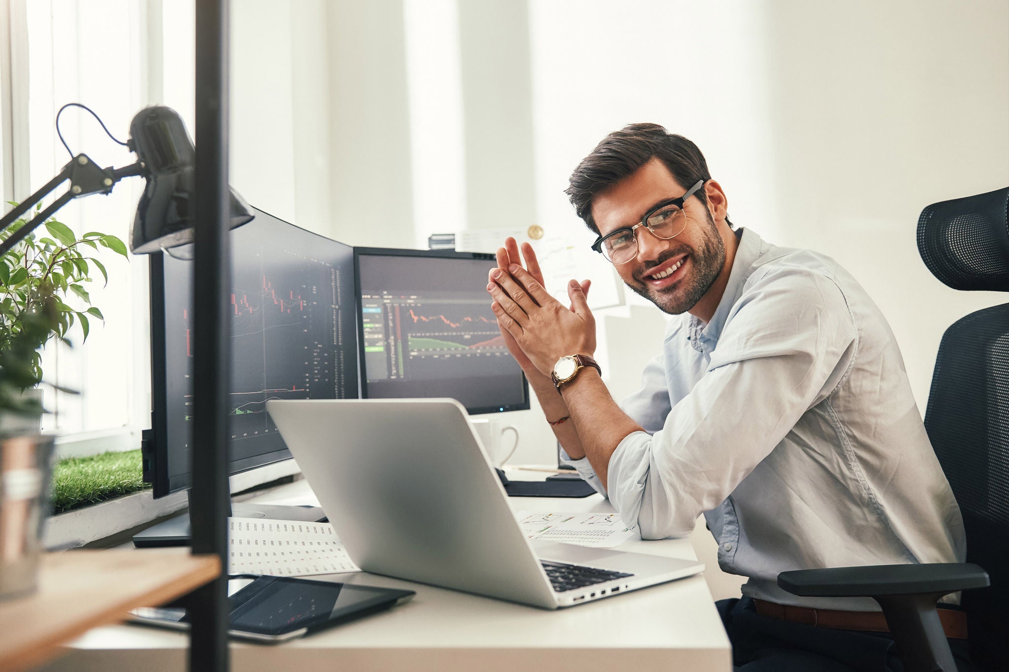 Young and successful. Happy bearded trader in formal wear and eyeglasses looking at camera and smiling while sitting in his modern office. Stock broker. Forex market. Trade concept