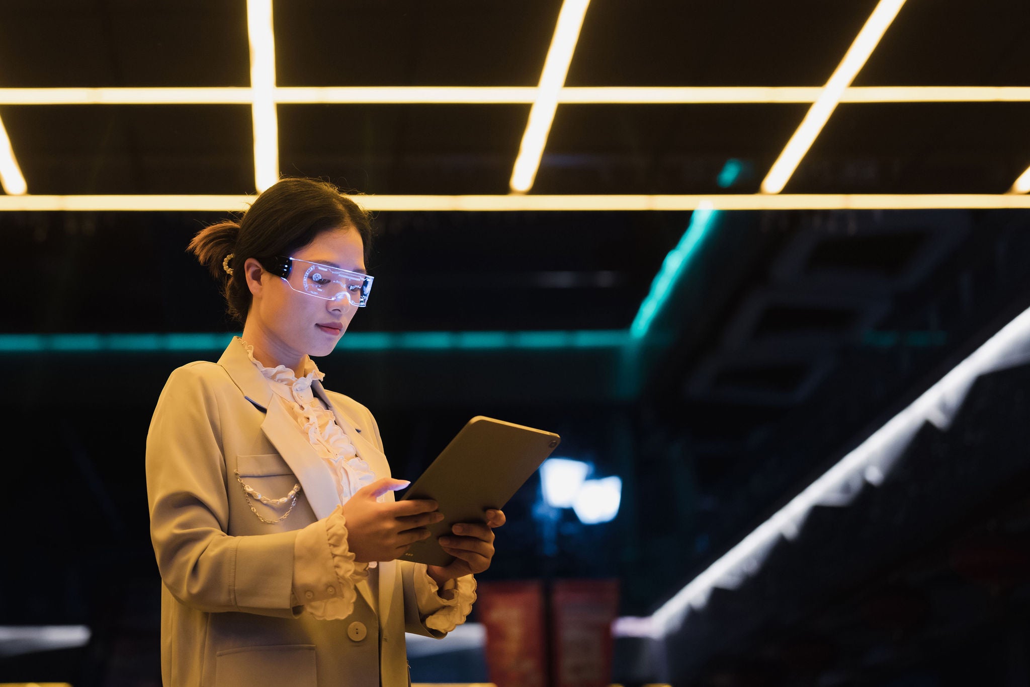 A young Asian business woman wearing glowing smart glasses uses a tablet in a city street at night. Concepts such as augmented reality, artificial intelligence, smart cities, metaverse, post-humanism are expressed.