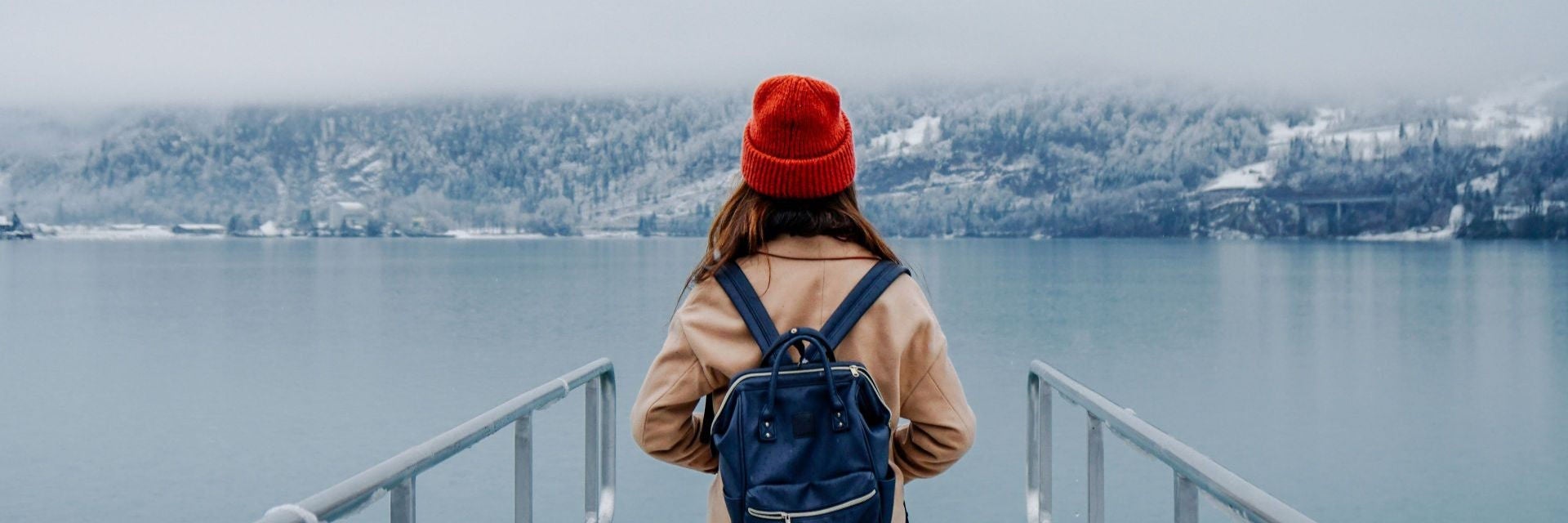 A person wearing a tan coat from behind, carrying a navy blue Anello-Style backpack with a wide opening and top handles