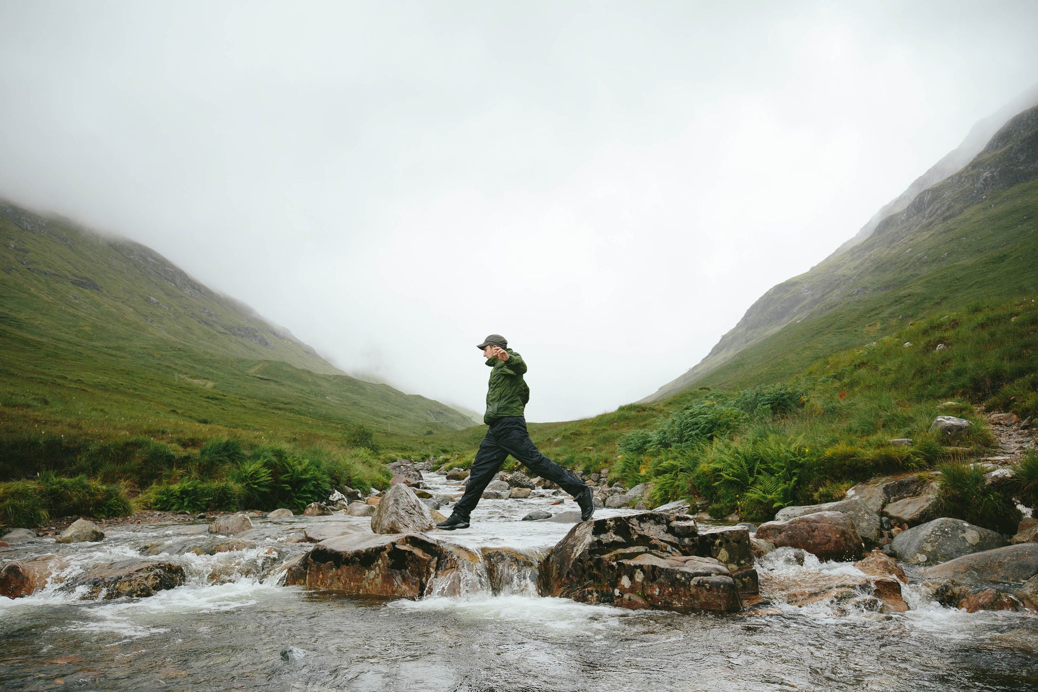 One man crossing river in lush green valley, Glencoe, Scotland