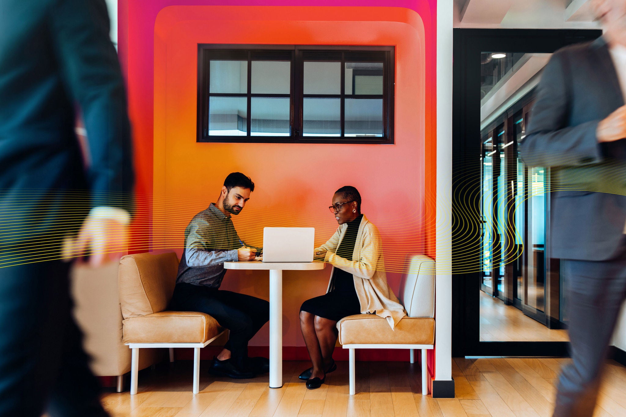 A man and woman sit together in a booth at a workplace, discussing work on a laptop. The space is cozy, designed with colorful partitions. Another professional is seen walking in the foreground.