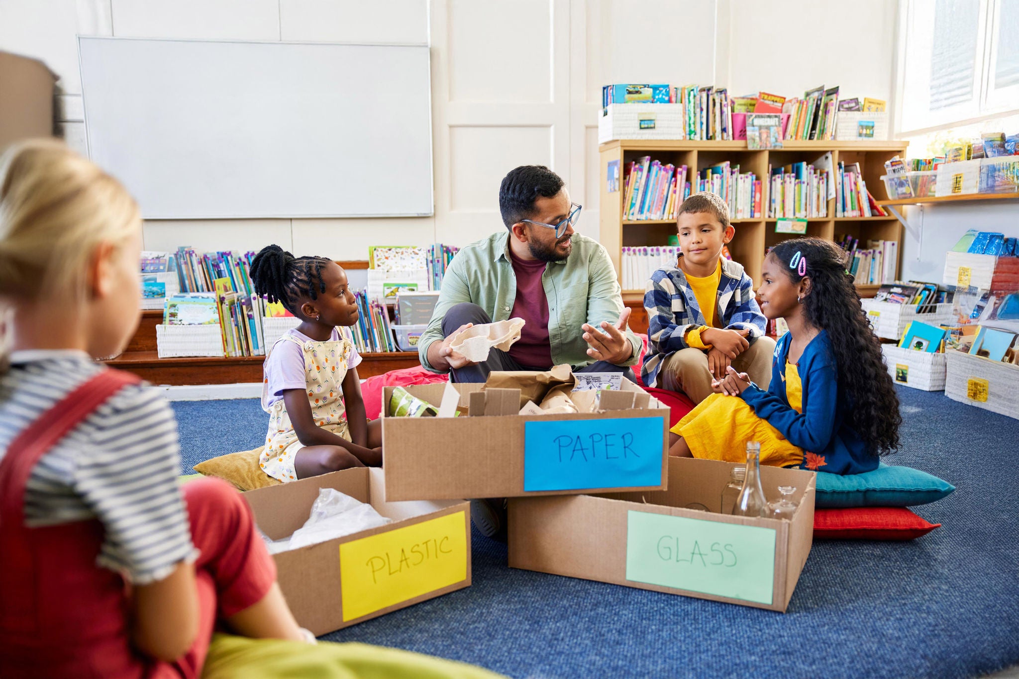Group of kids with male teacher organizing paper, plastic and glass in cardboard boxes in classroom to throw them away separately. Teacher educates the elementary class in separating waste: paper, plastic and glass for recycling. Multiethnic children participate in the separation of plastic, paper and glass waste to save the planet from pollution.