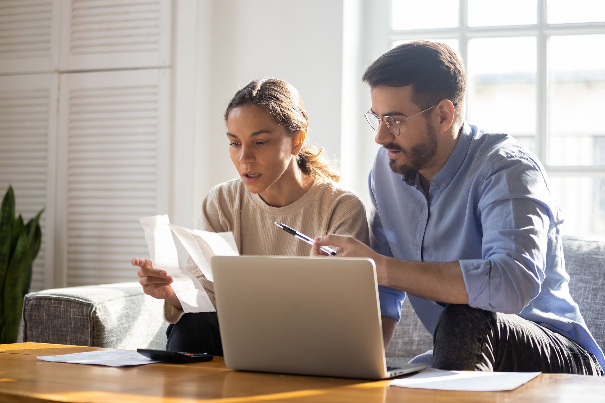 young couple sitting on a couch, reviewing papers using laptop