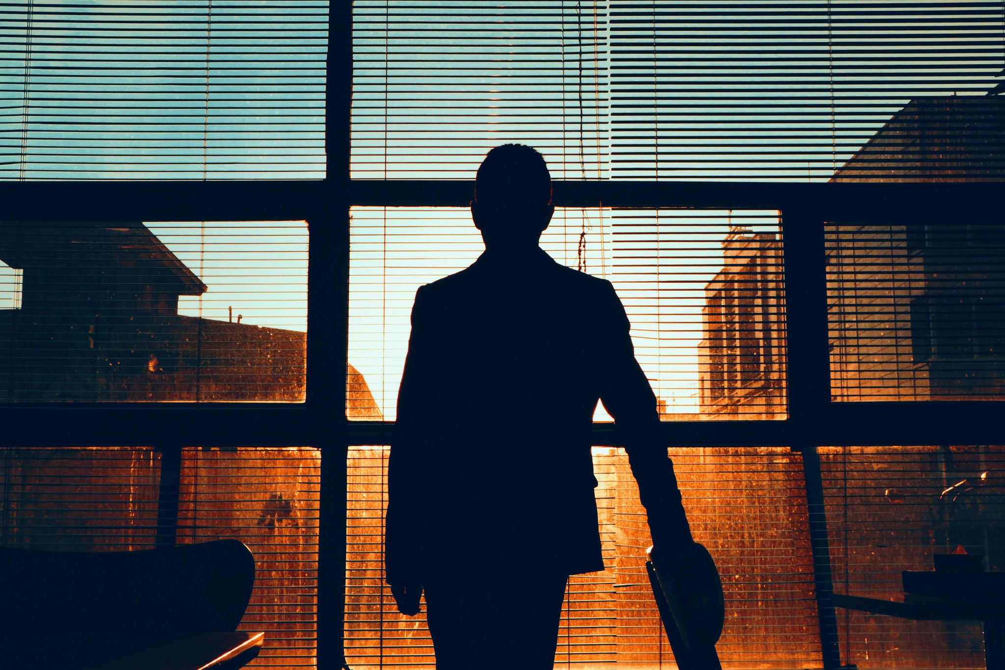 Silhouette of a person standing in front of large windows with blinds, looking at buildings during sunset