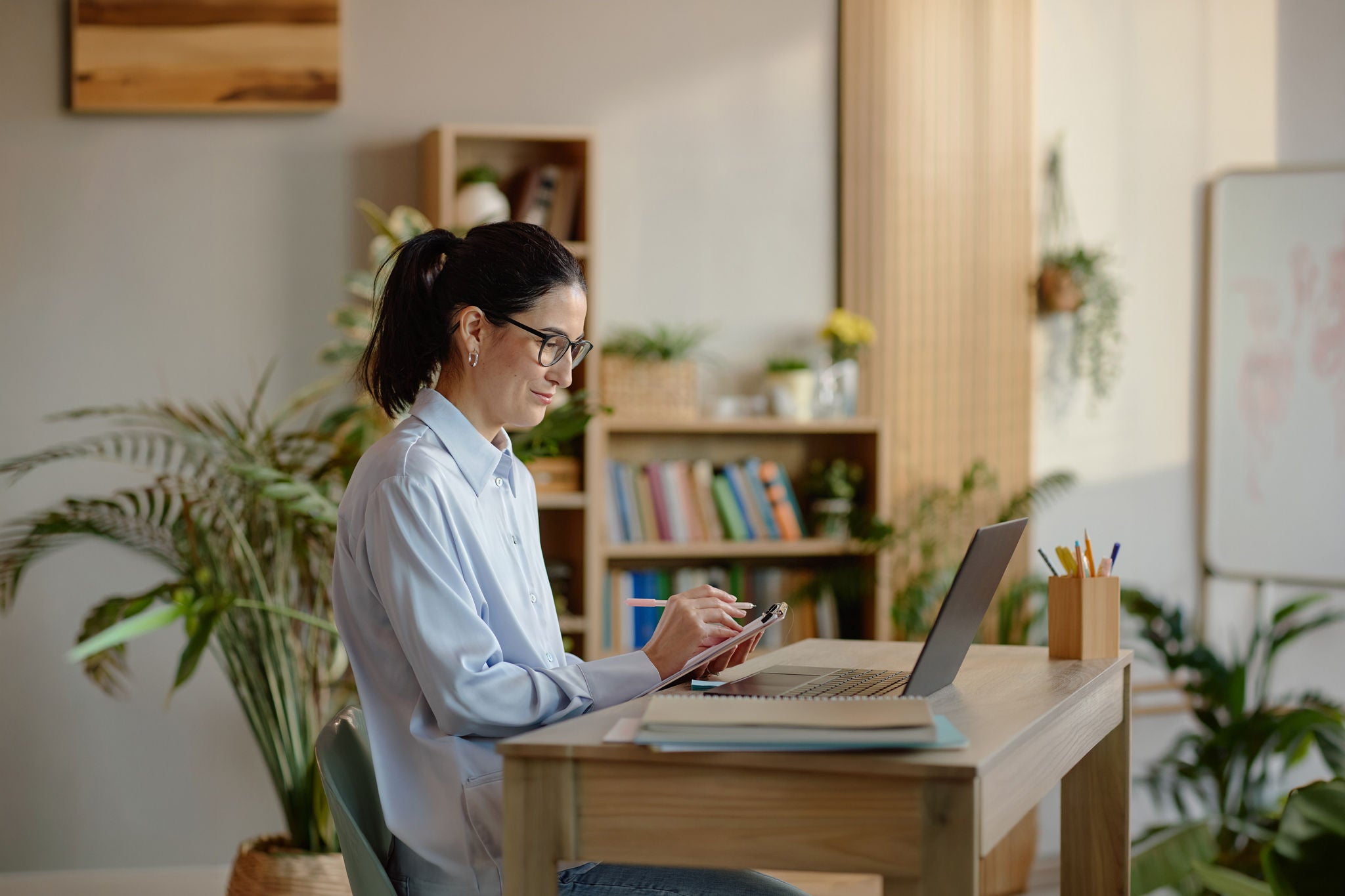 Caucasian young adult woman sitting at desk writing on clipboard while looking at laptop screen, wearing glasses, working in office setting with bookshelves and plants in background