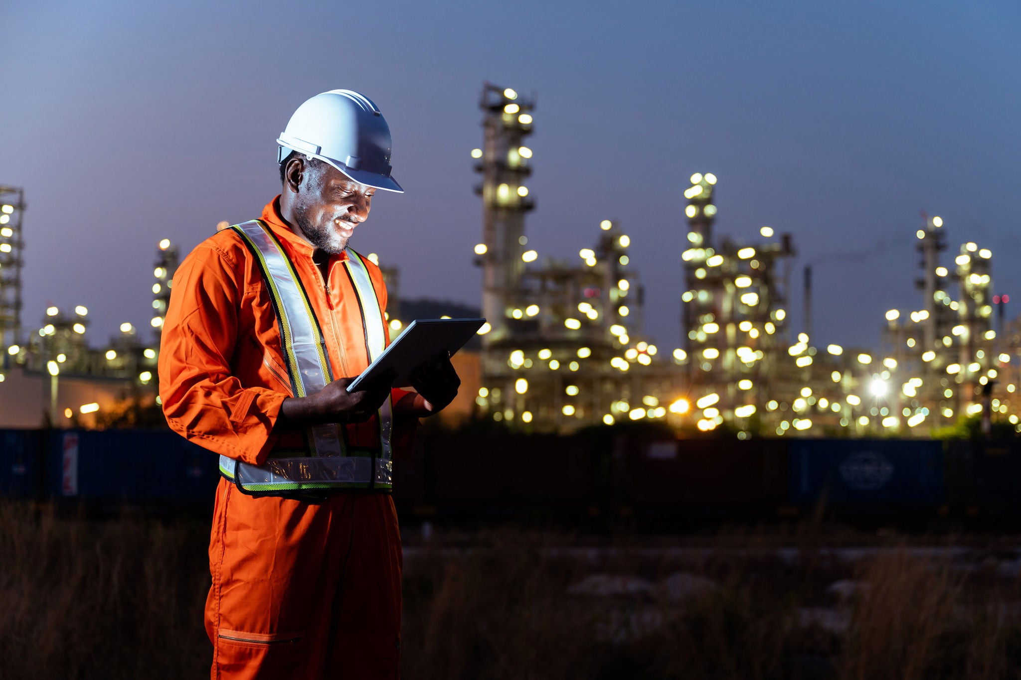 Construction worker using a tablet at an industrial site at night with illuminated facilities in the background