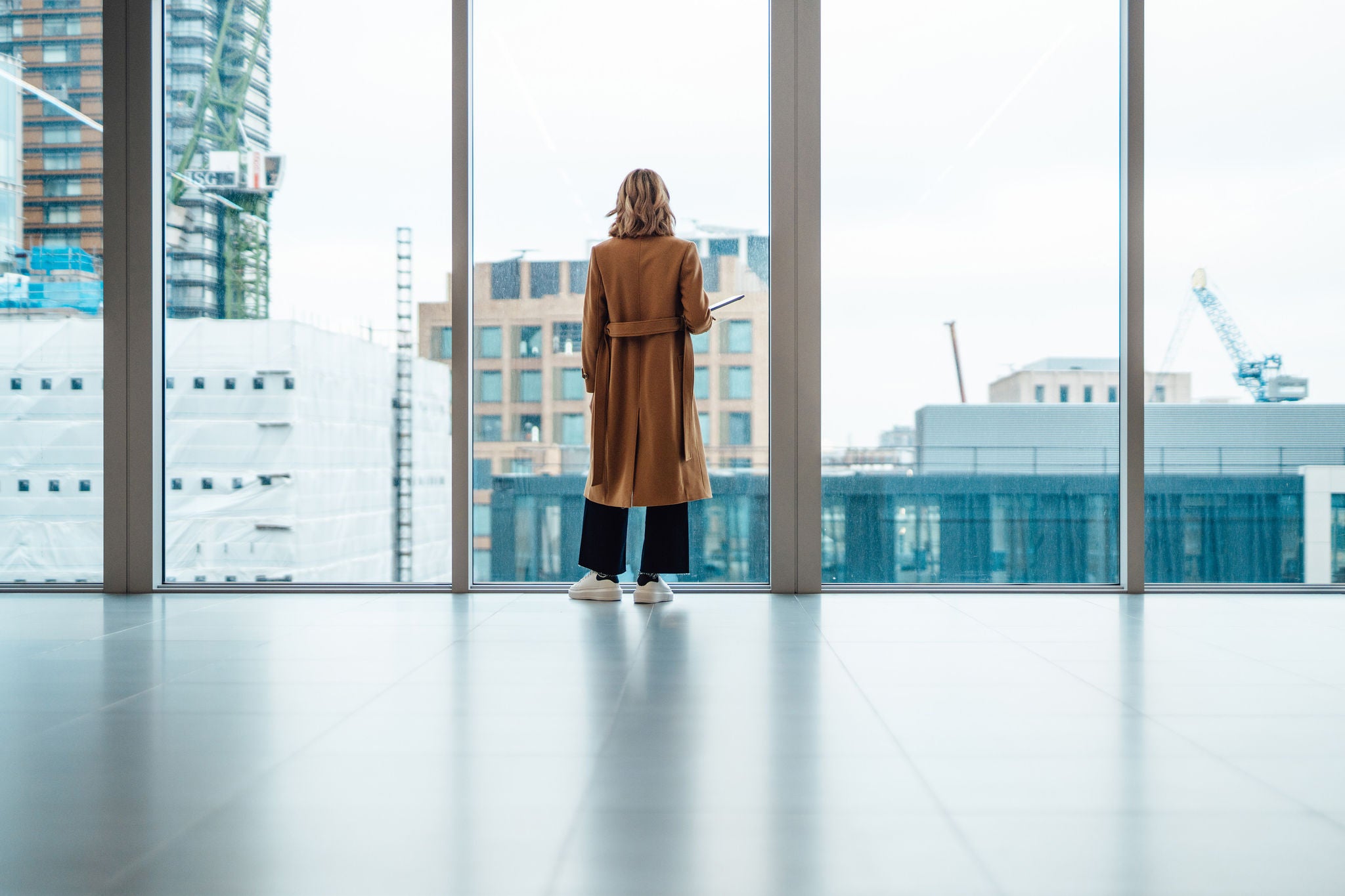 Back view of businesswoman standing and looking through window in conference room. She is thinking business strategy and making new business plan. Holding a laptop on her arm. Transgender female pursuing her dreams and successful career.