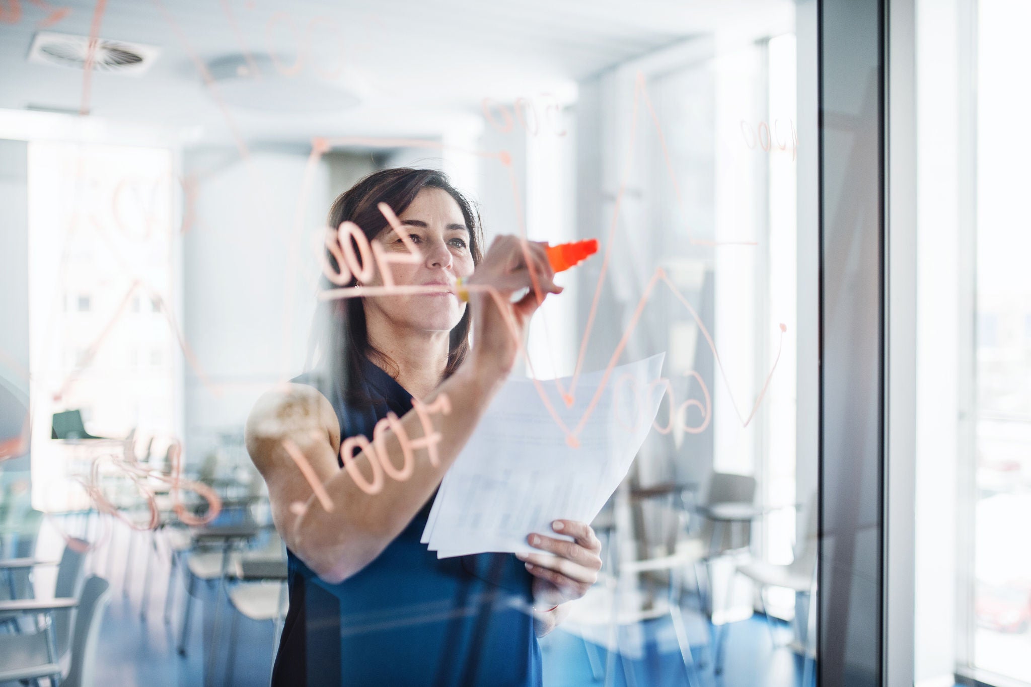 Woman writing notes on a glass board while holding papers in a modern office