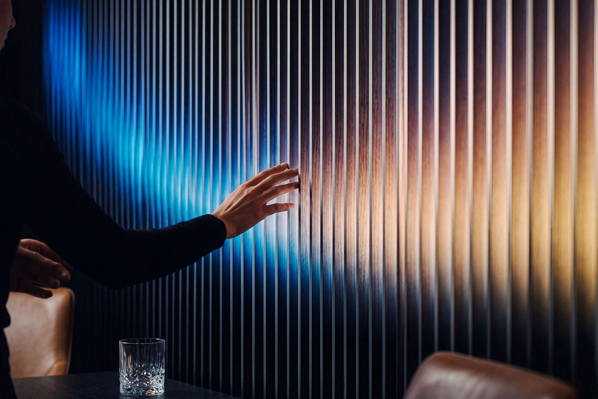 A human hand touching a sleek modern vertical slatted wall illuminated with vibrant colorful light.