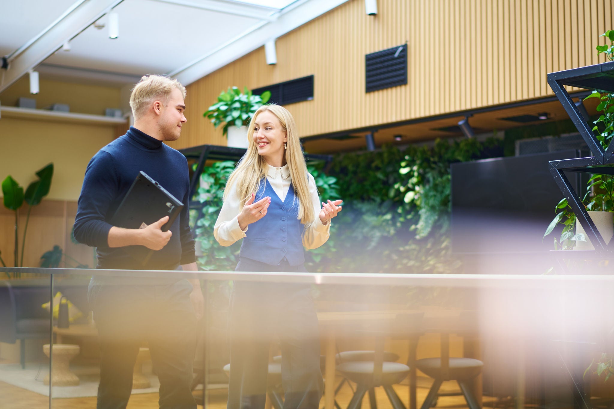Two colleagues walking and discussing in a modern office with indoor greenery and open collaborative spaces.