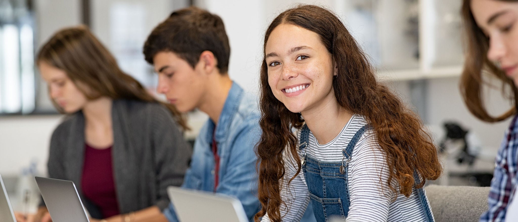 Happy young woman working on laptop and looking at camera in classroom. Portrait of smiling university student in library use computer for a research. Satisfied college student looking at camera while sitting in a row with classmates studying together.