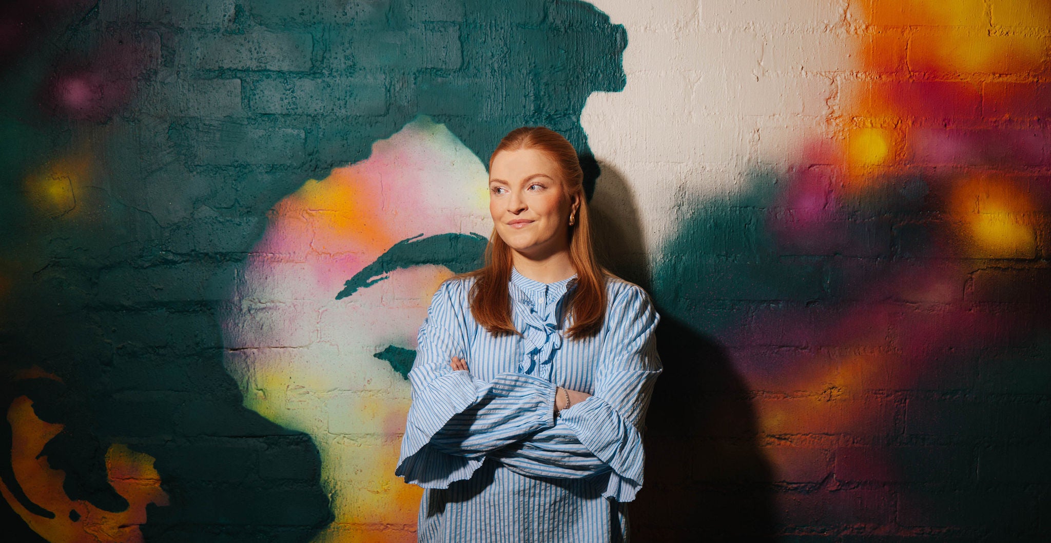 Woman standing with folded arms in front of a colorful painted brick wall.