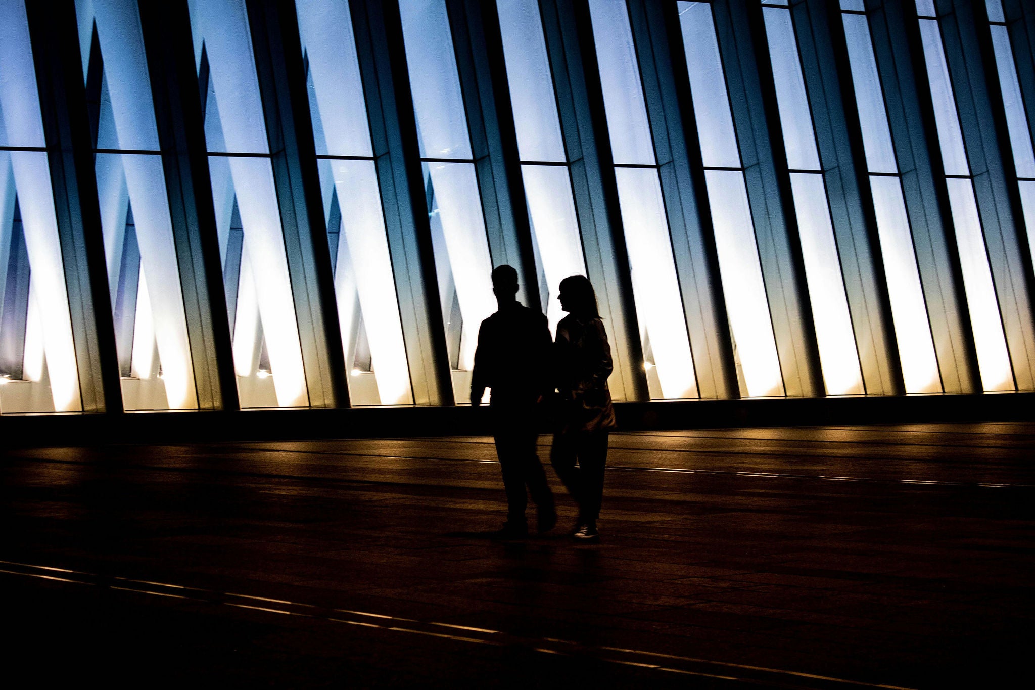 Two people walking in front of a modern illuminated glass facade at dusk
