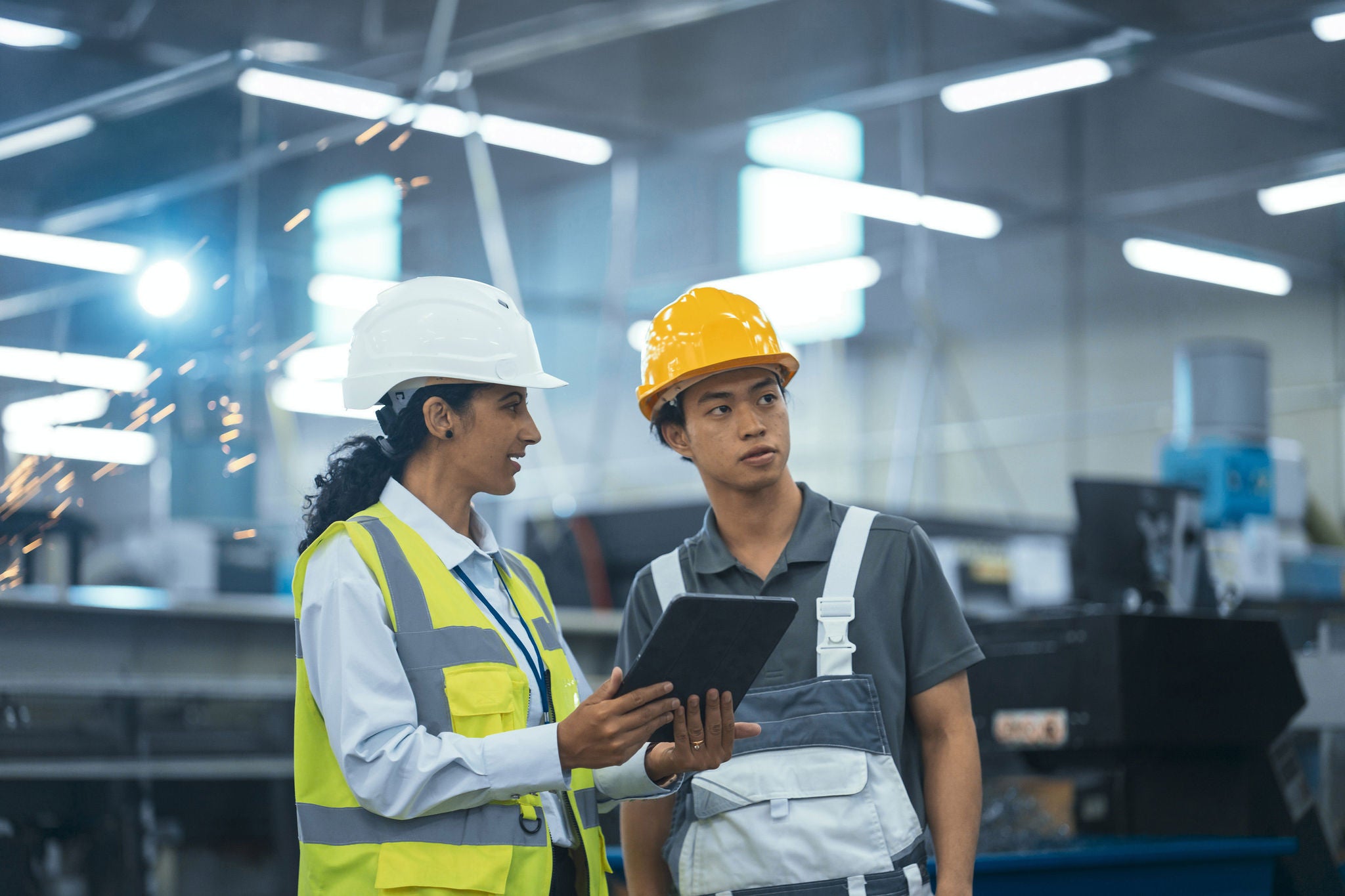 Young, diverse factory team in hard hats as an engineer shows tablet data to a technician amid sparks.