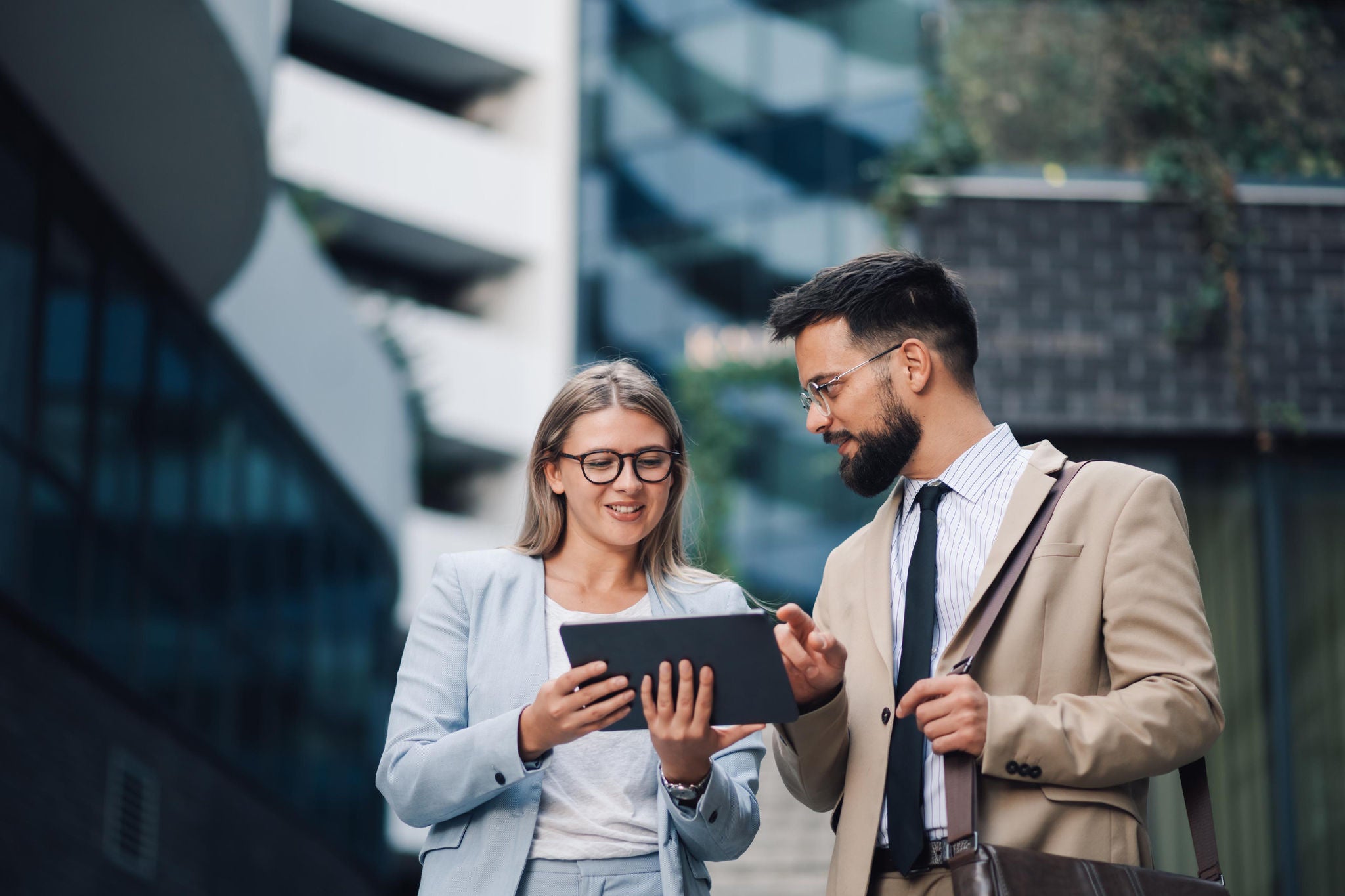 Two business partners are discussing business strategy while reviewing project data using a digital tablet in front of an office building