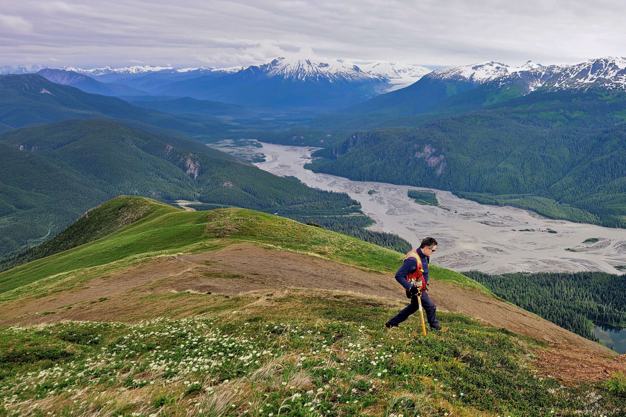 Person hiking on a mountain ridge overlooking a wide valley and snow‑covered peaks