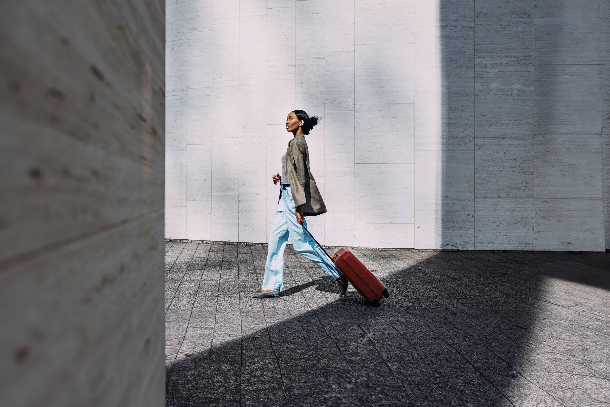 Confident woman walking with red suitcase against a modern urban wall