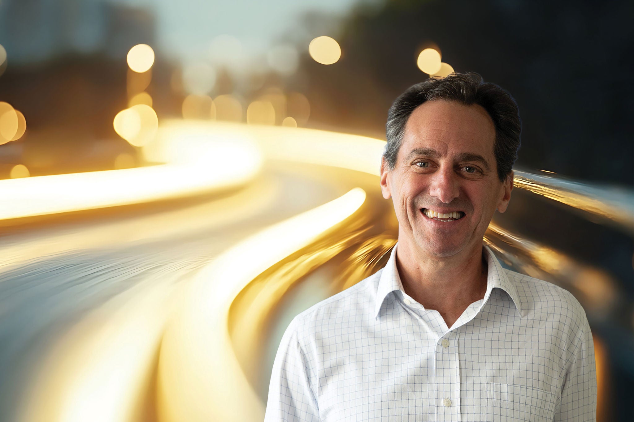 Smiling man in a light-colored shirt standing beside flowing golden light trails on a dark background.