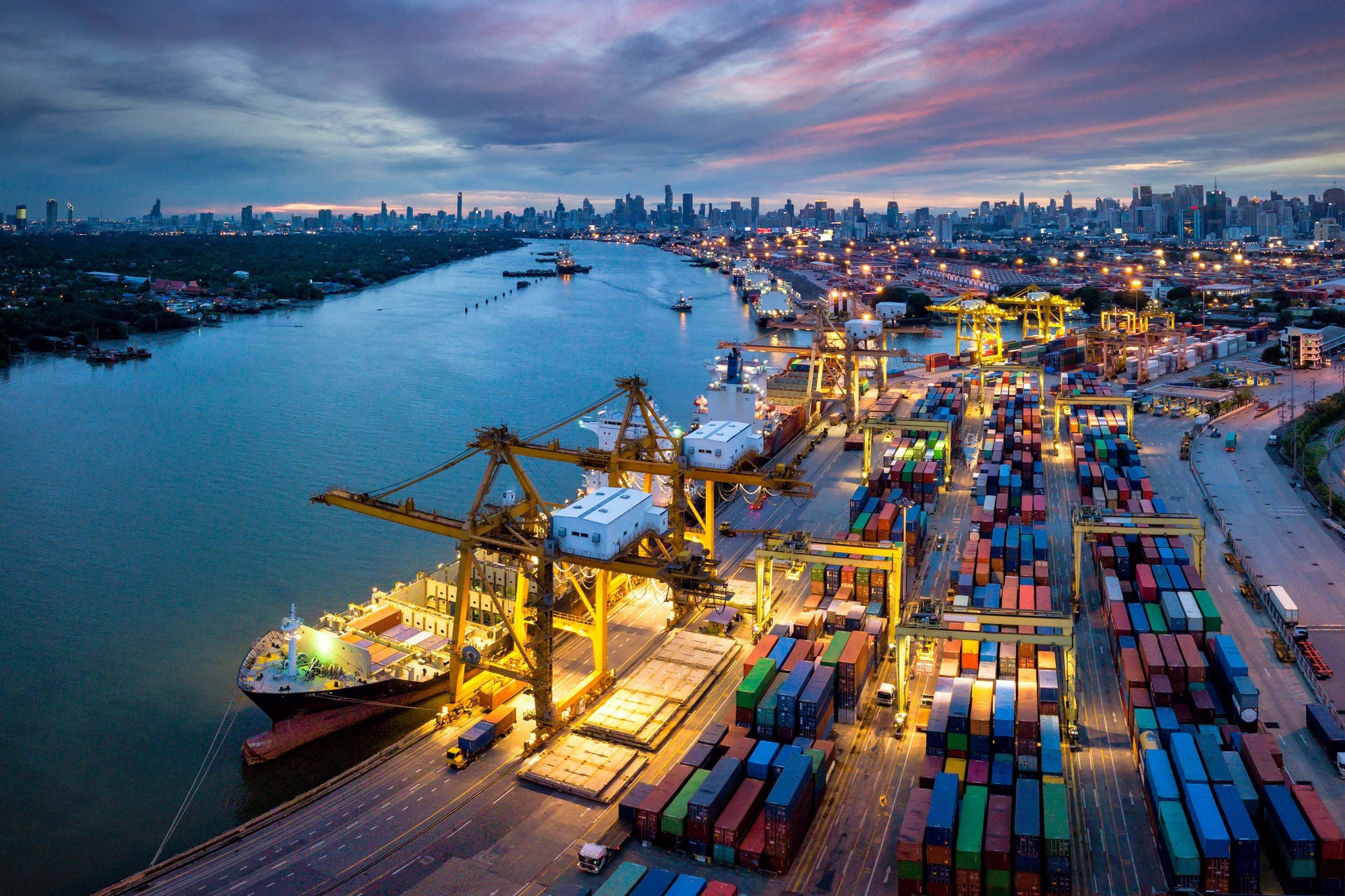 Moderner Containerhafen mit Kränen, Containerschiffen und beleuchteter Skyline am Fluss.