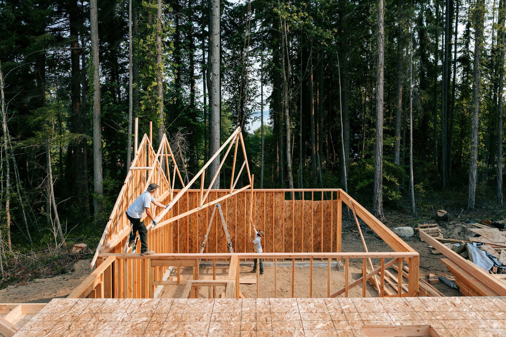 Workers build the frame of a new house project.  Bare plywood and beams as it is framed up from the foundation.  High lumber costs have affected the building process.  Shot in Washington state, USA.  High angle drone point of view.