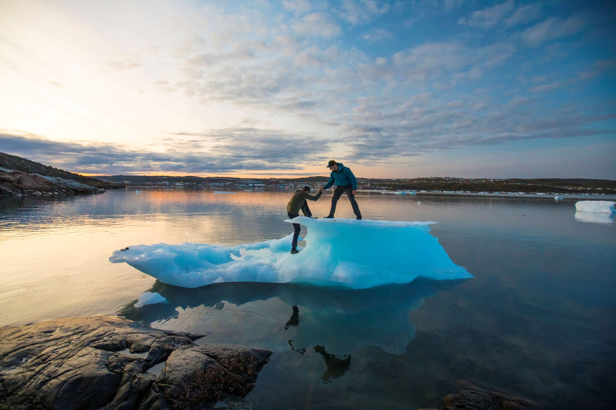 Two people standing on a small iceberg in calm water during sunset.