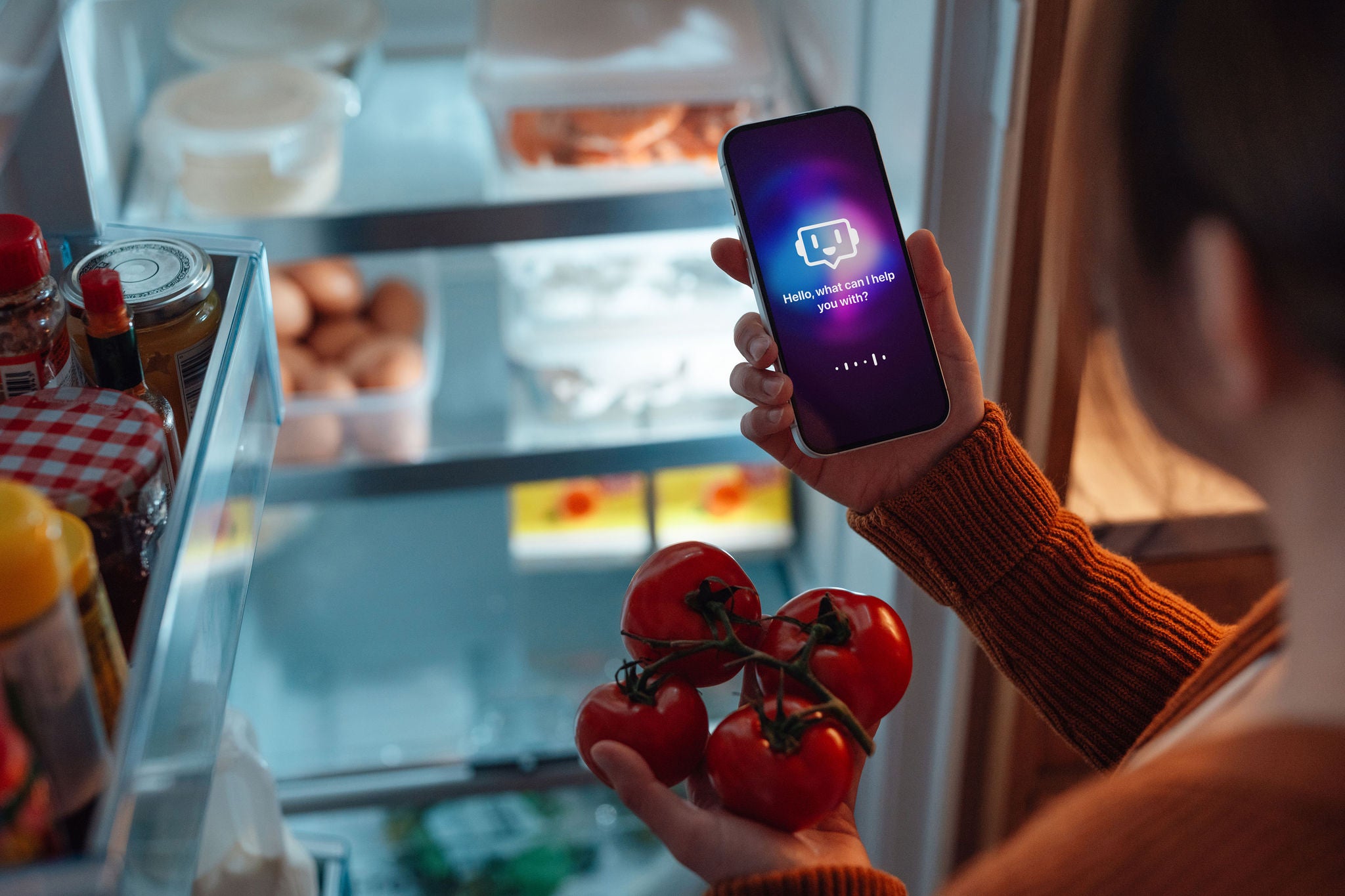 Person using a smartphone AI assistant while selecting tomatoes from an open refrigerator.