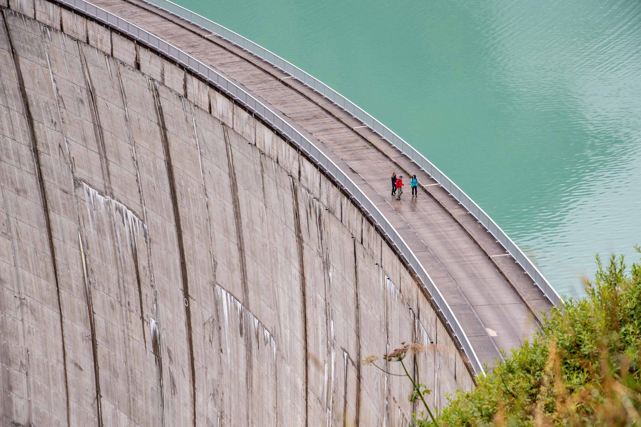 Drei Personen gehen über Staumauer neben türkisfarbenem Wasser