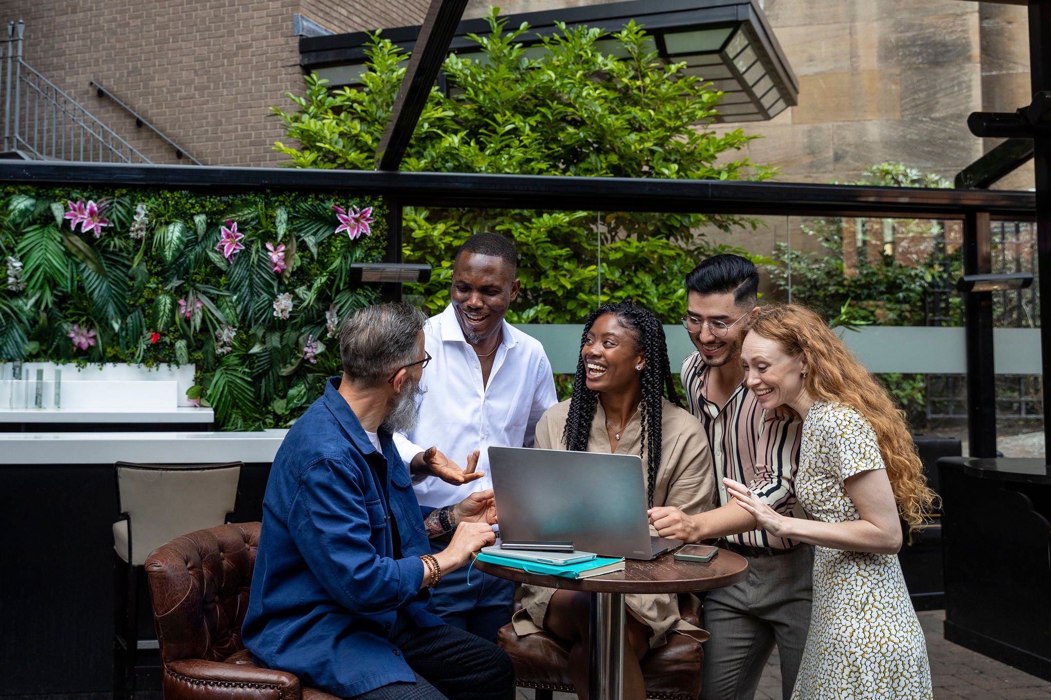 A wide-view shot of a mixed ethnic group of business professionals talking together while sitting outside on a patio. They are having a meeting on a video call with one of their boss in the North East of England.