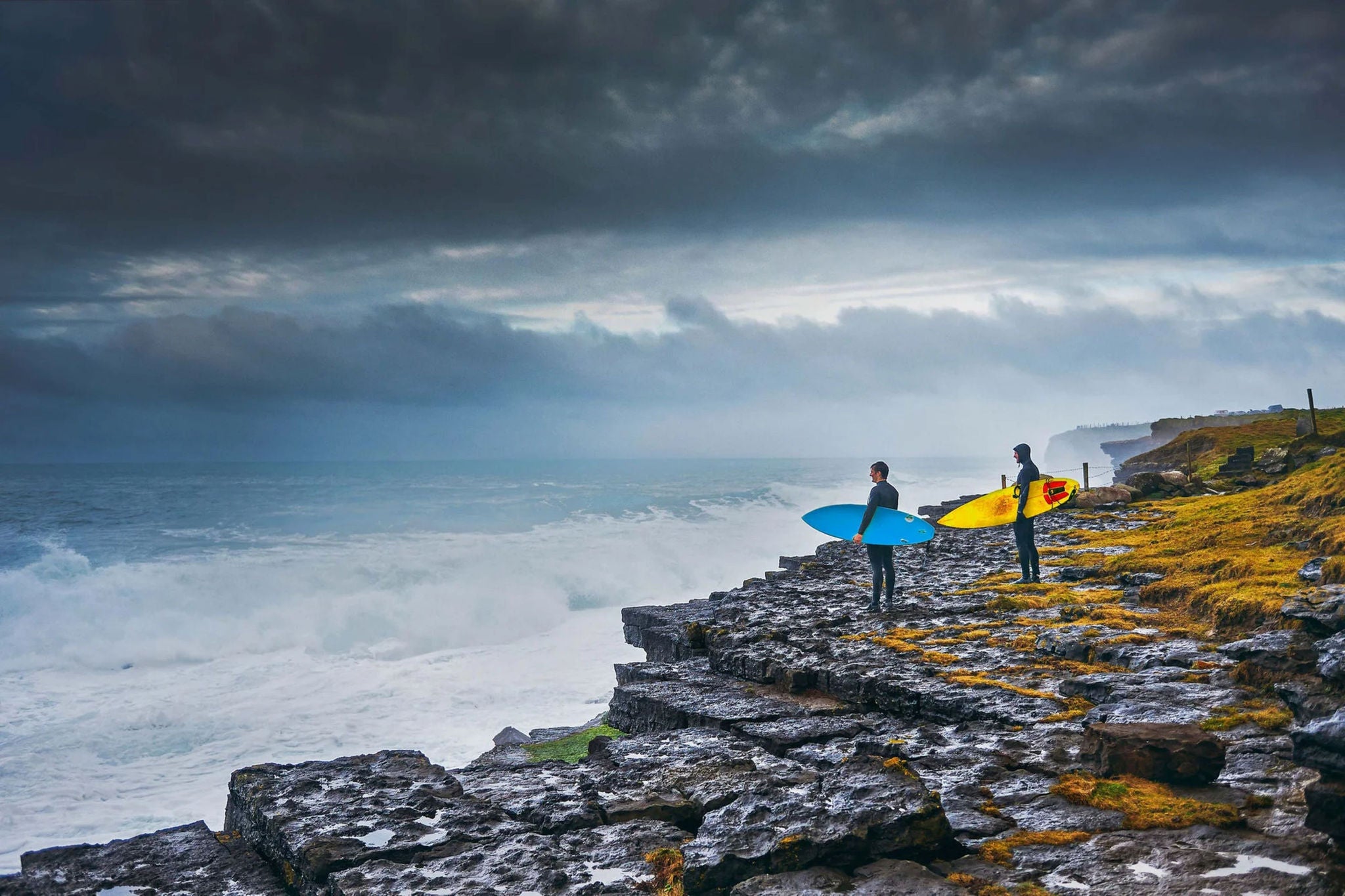 Surfeurs avec planches sur falaise rocheuse face à l’océan agité.