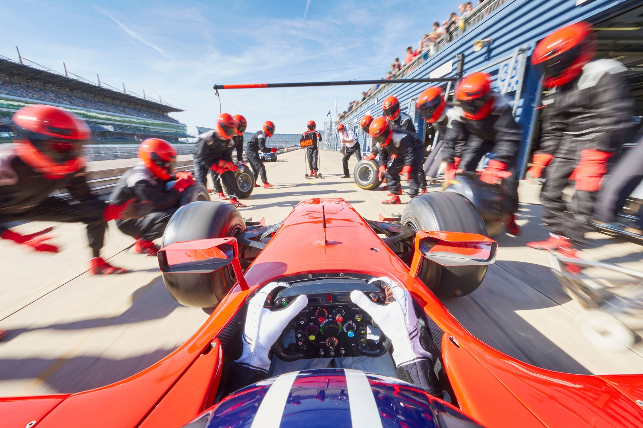 Driver's view of a red Formula One car during a high-speed pit stop as crew members change tires.