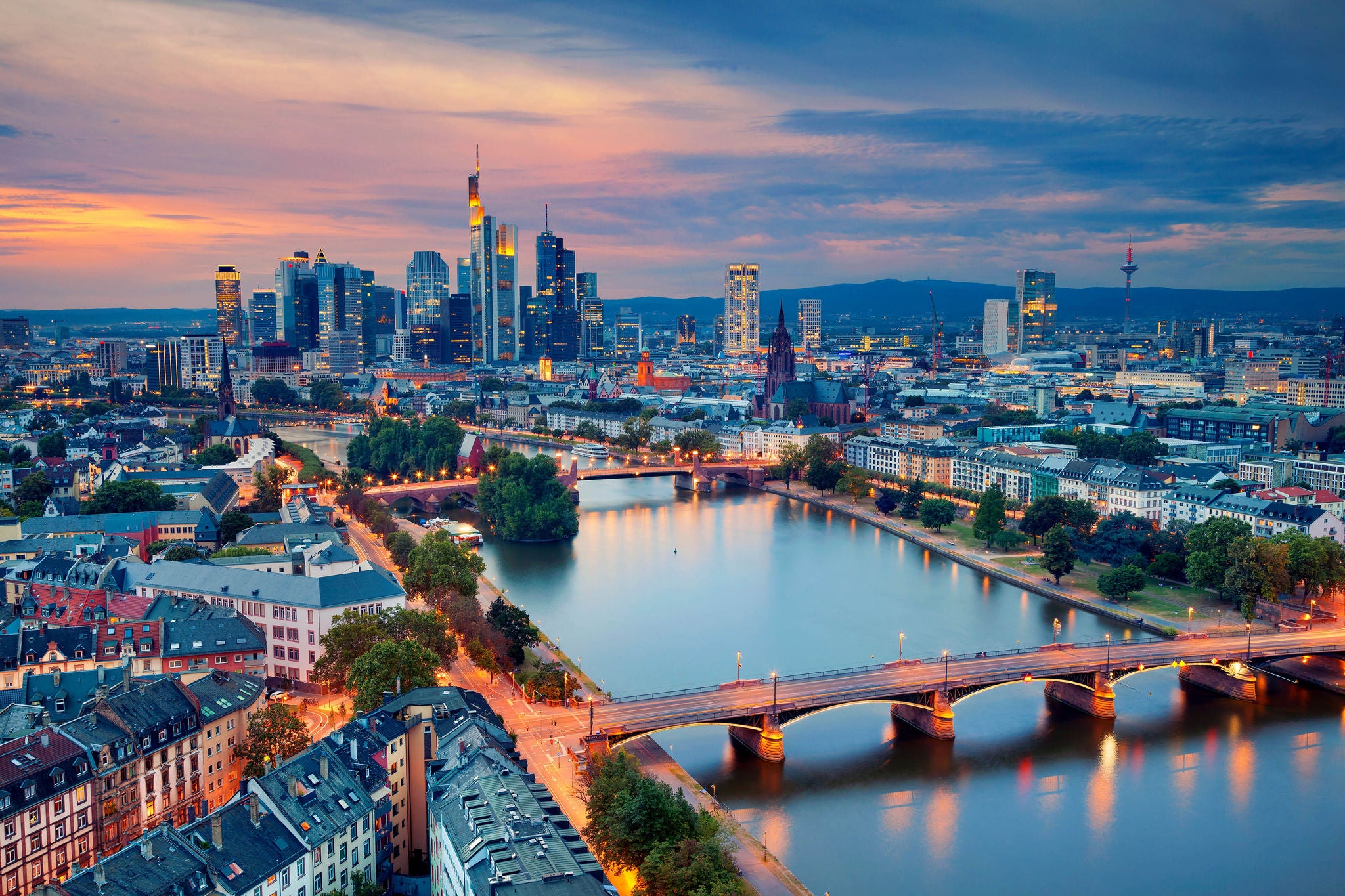 Panoramablick auf Frankfurt mit Mainbrücke und leuchtenden Gebäuden im warmen Abendlicht.