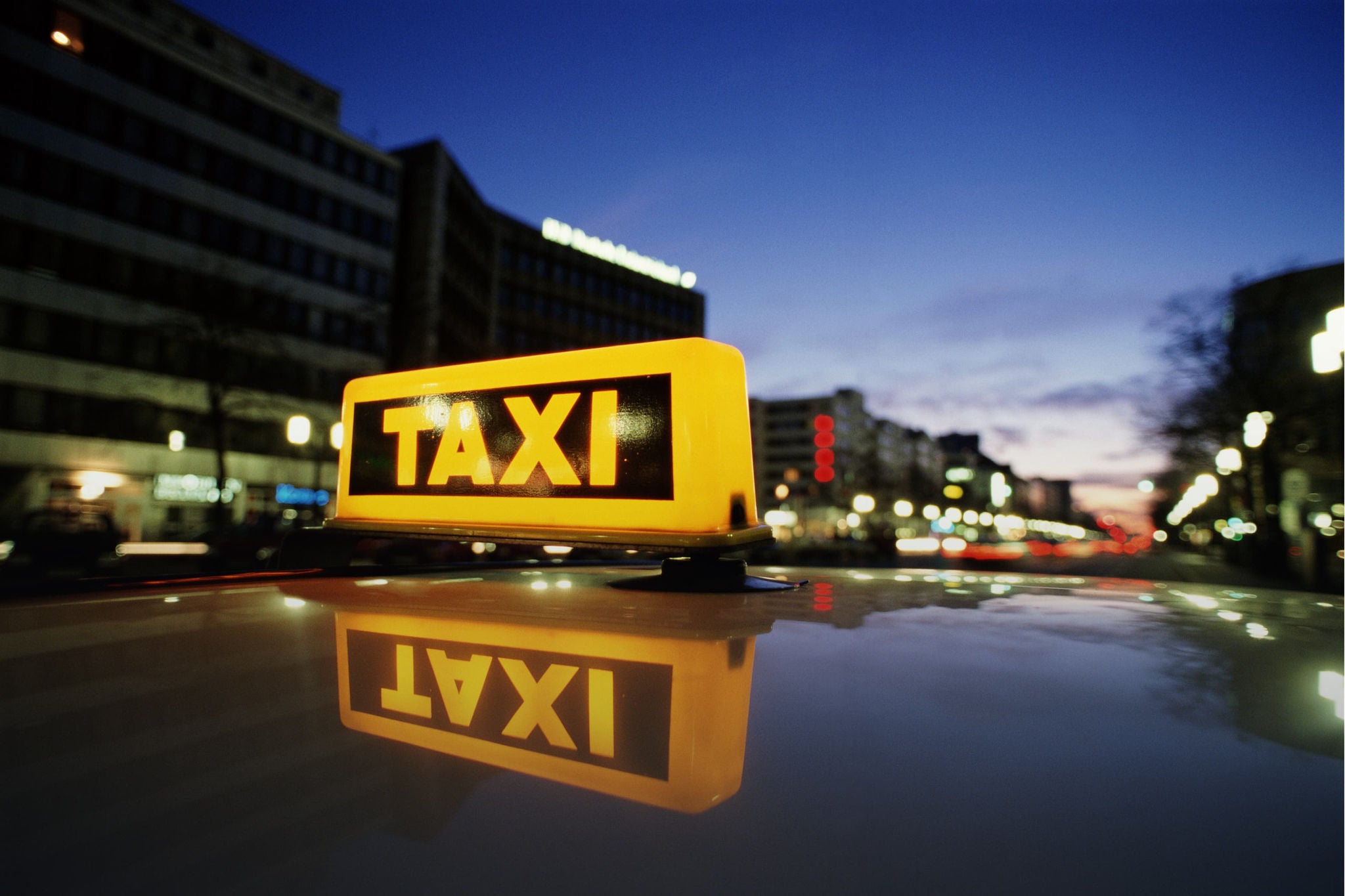 Ein beleuchtetes Taxischild spiegelt sich auf dem Autodach in einer nächtlichen Stadt.