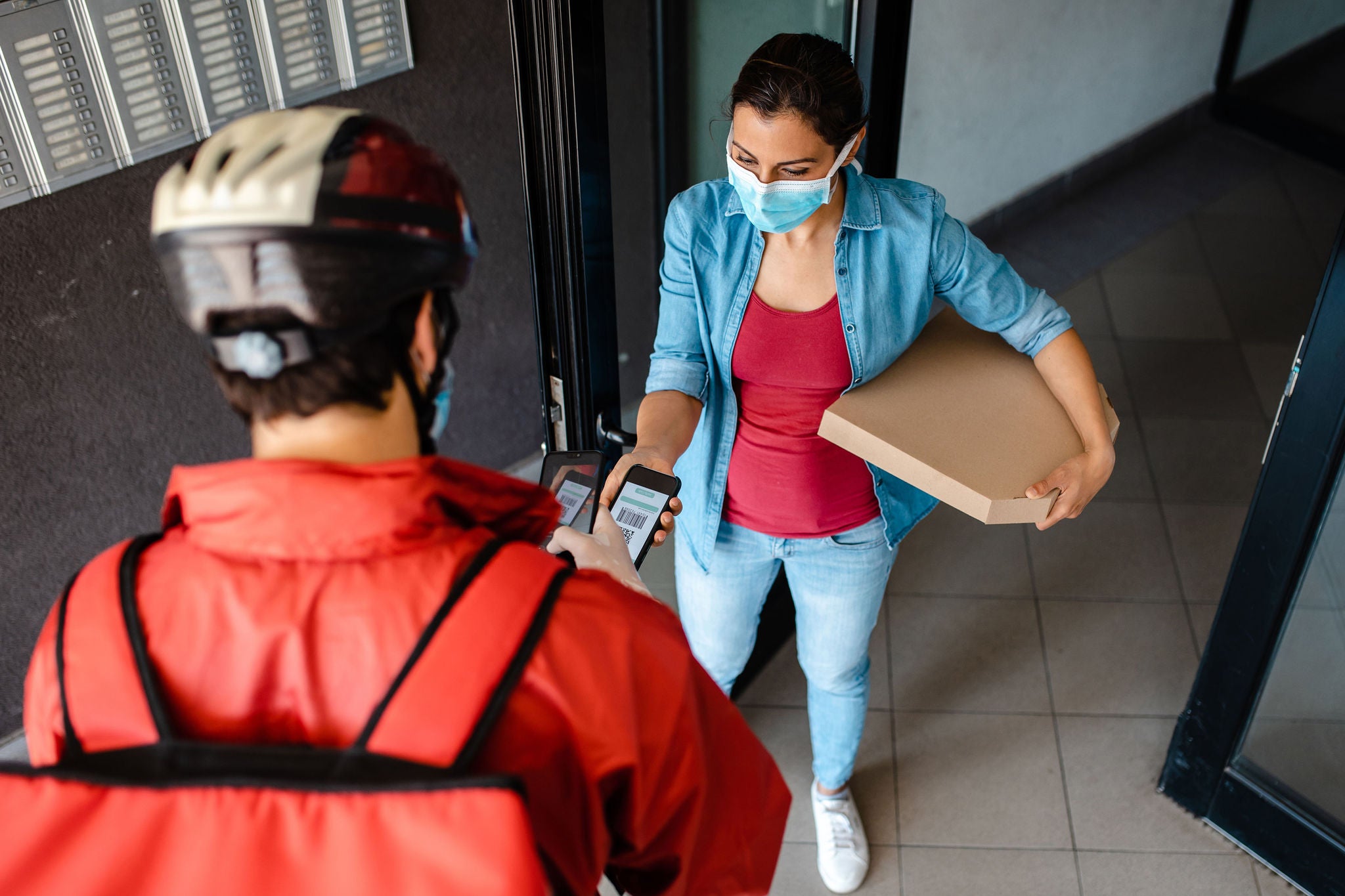 Delivery guy deliver a pizza to a customer in front of the residential building, and she paying via mobile app so they avoid a contact during corona virus pandemic