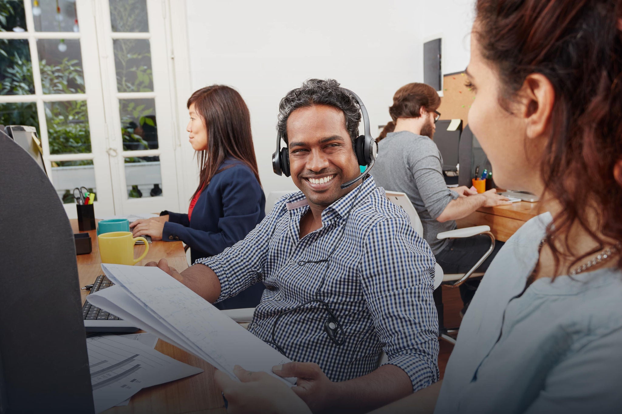 A smiling young woman with headphones and folders, standing outdoors on a college campus.