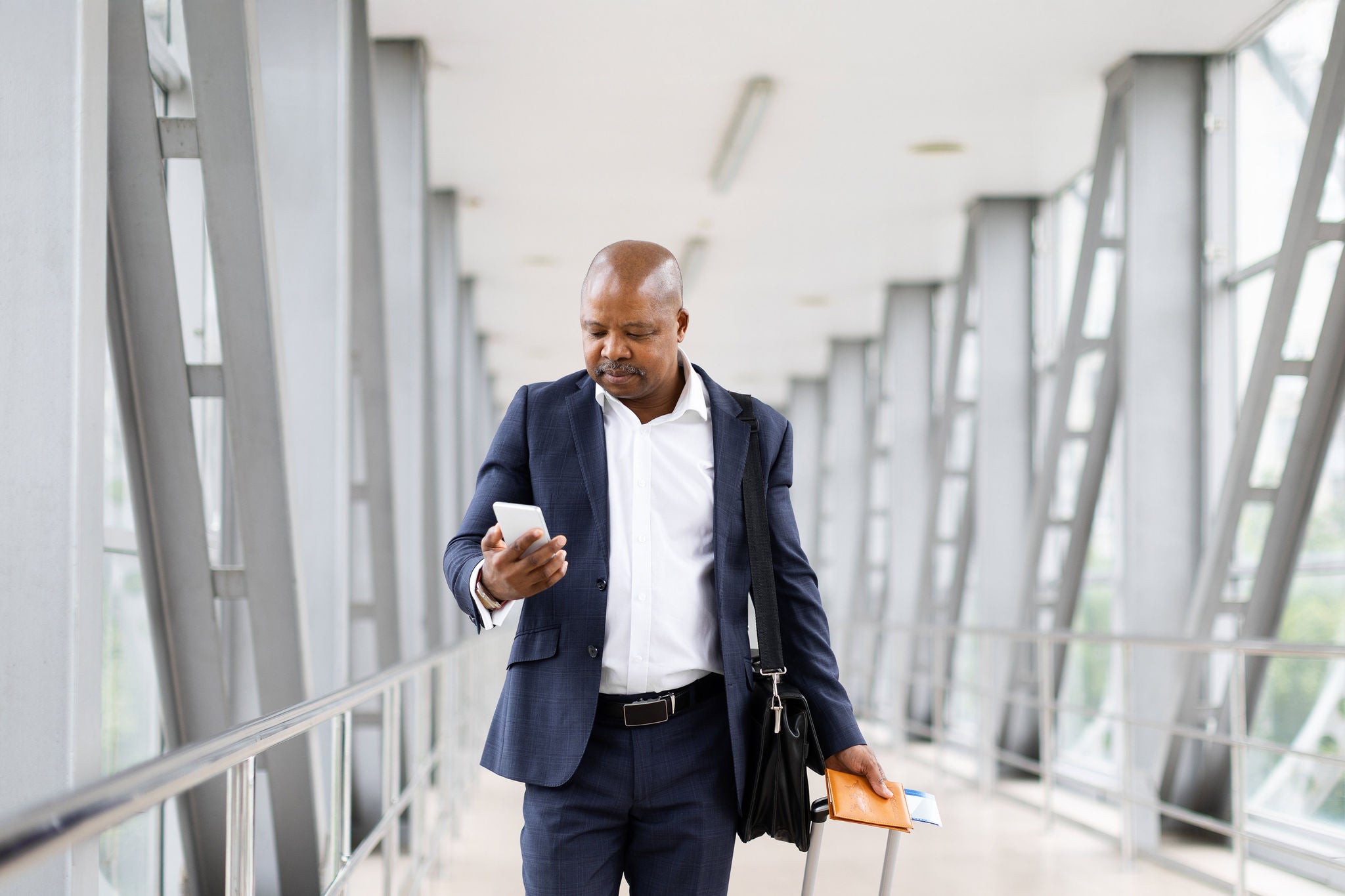 Focused African American businessman walking in airport terminal, checking smartphone and holding passport with suitcase, preparing for international trip