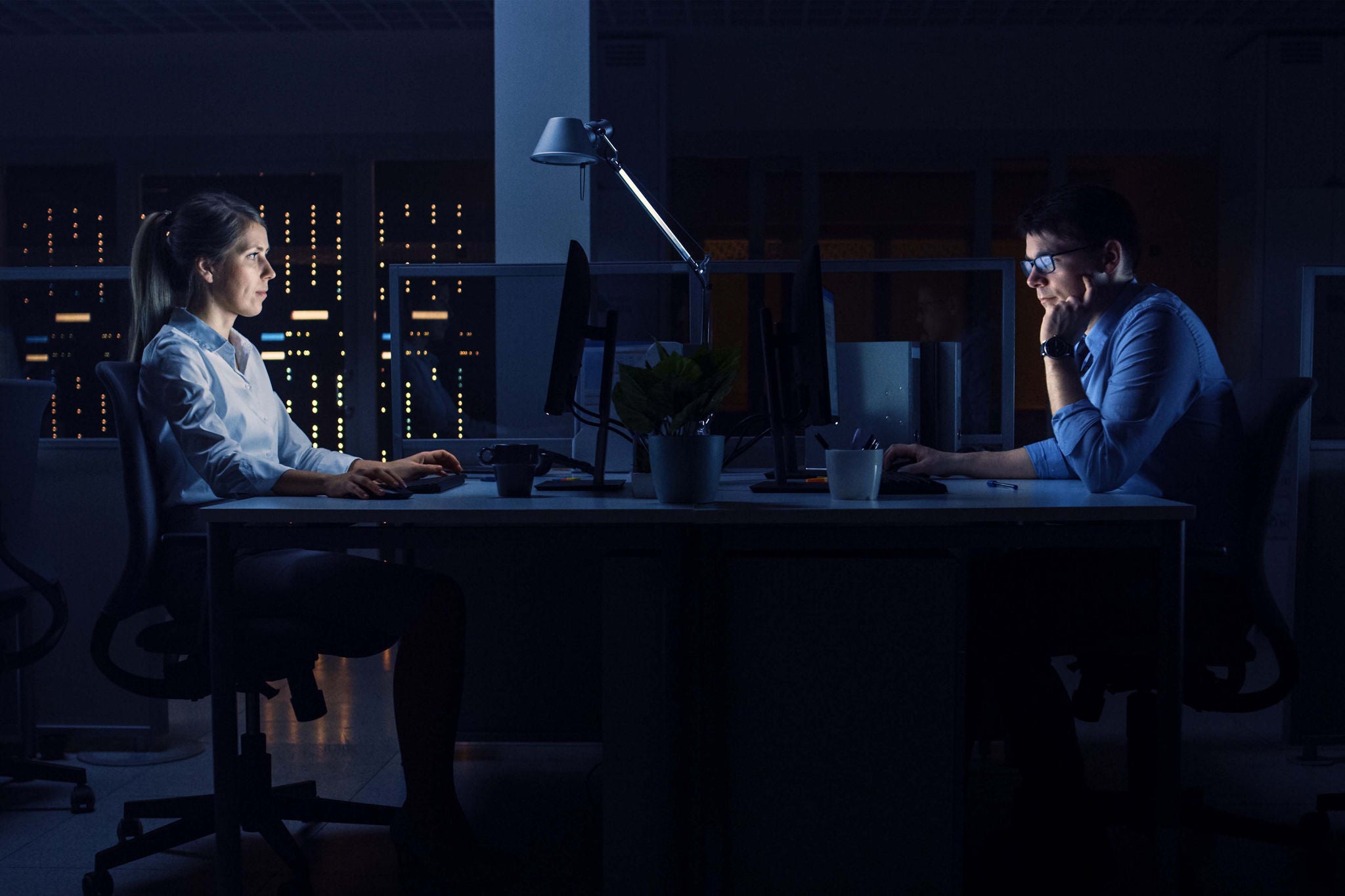  Two professionals work late in a dimly lit office with computers and city lights in the background.