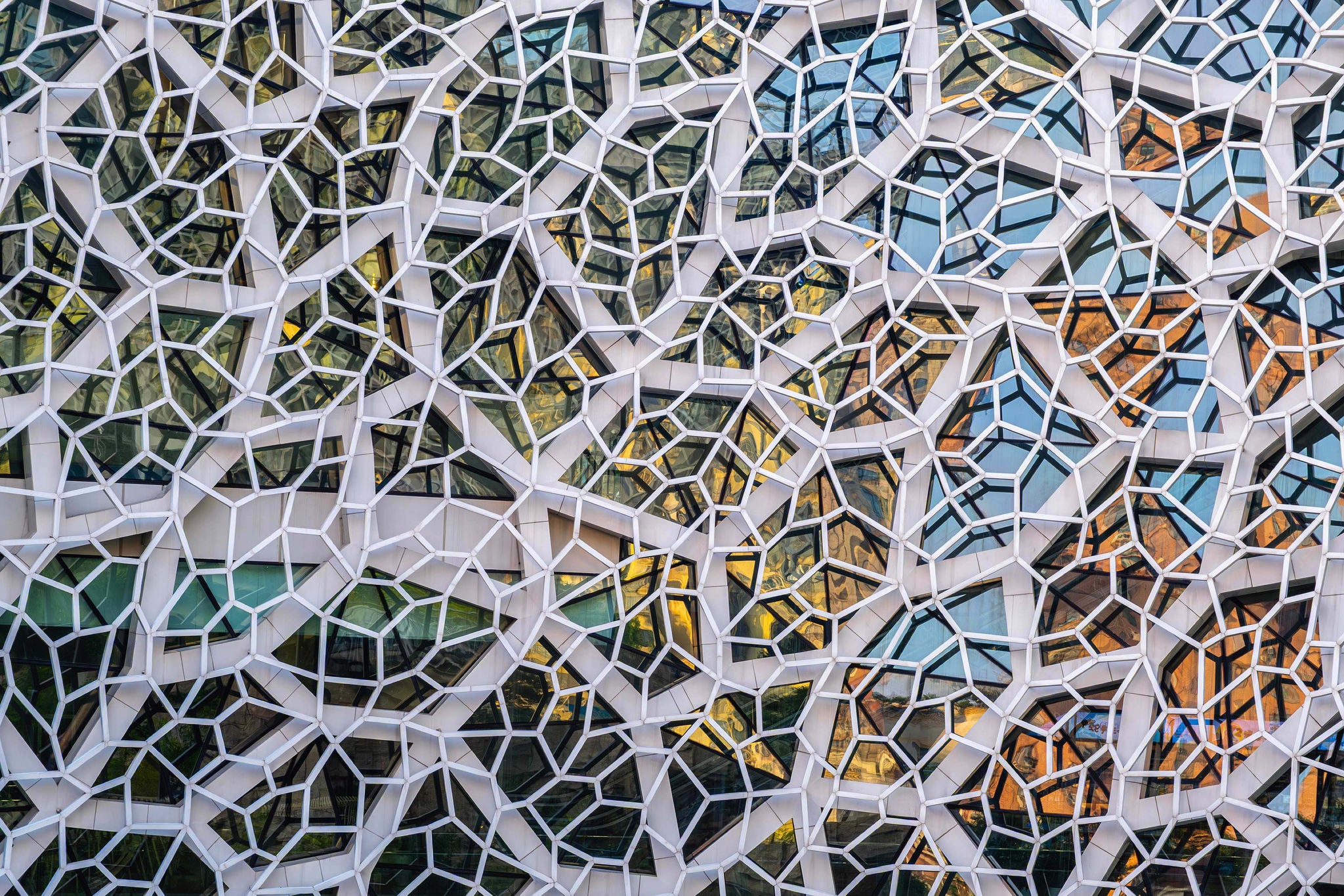 Modern facade with white hexagonal and irregular frames enclosing glass panels in a geometric mosaic pattern.