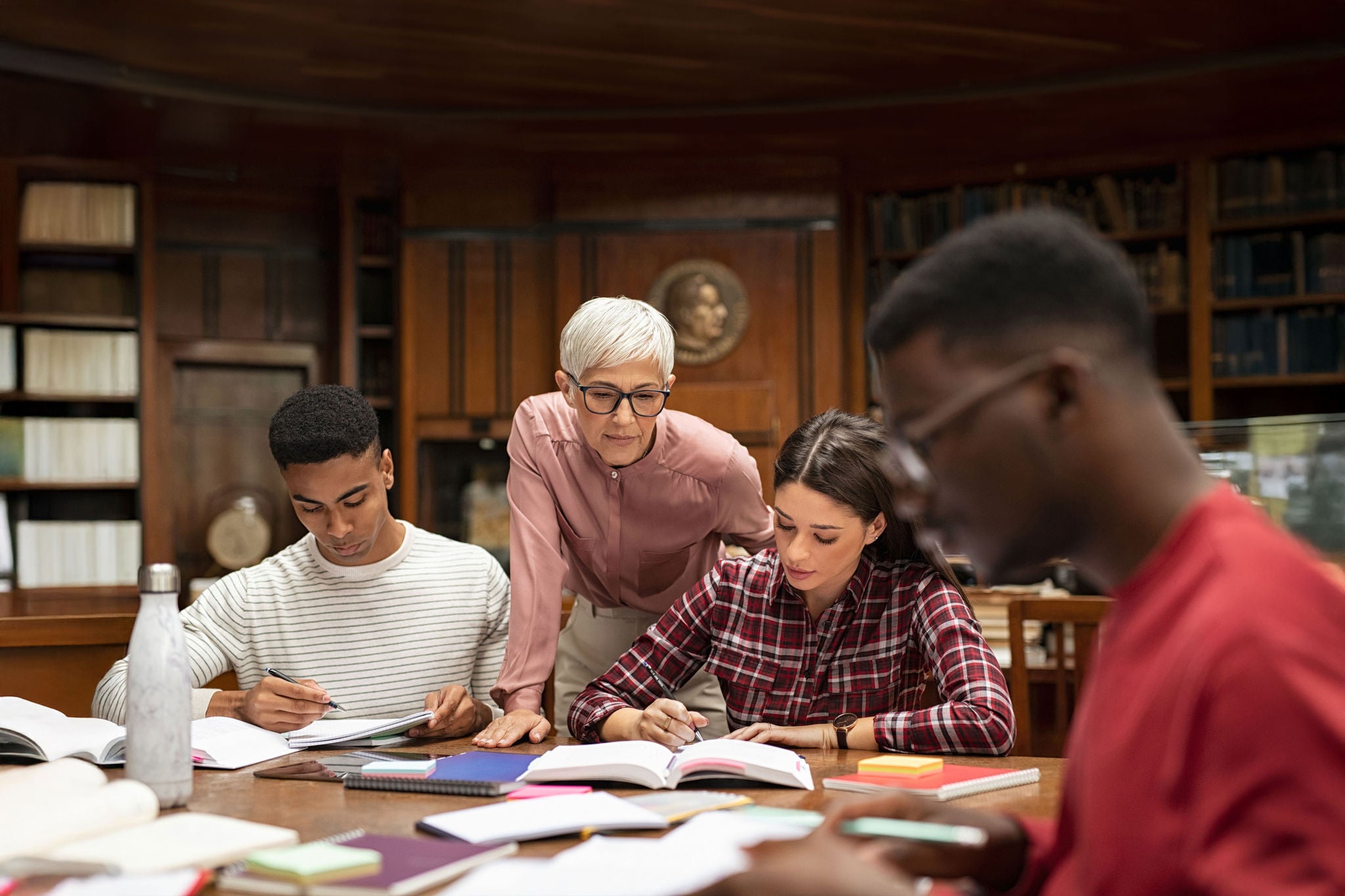 Senior woman teacher working with college students in library. Graduates consulting with their lecturer informally during a break in library. Group of students studying with their professor.