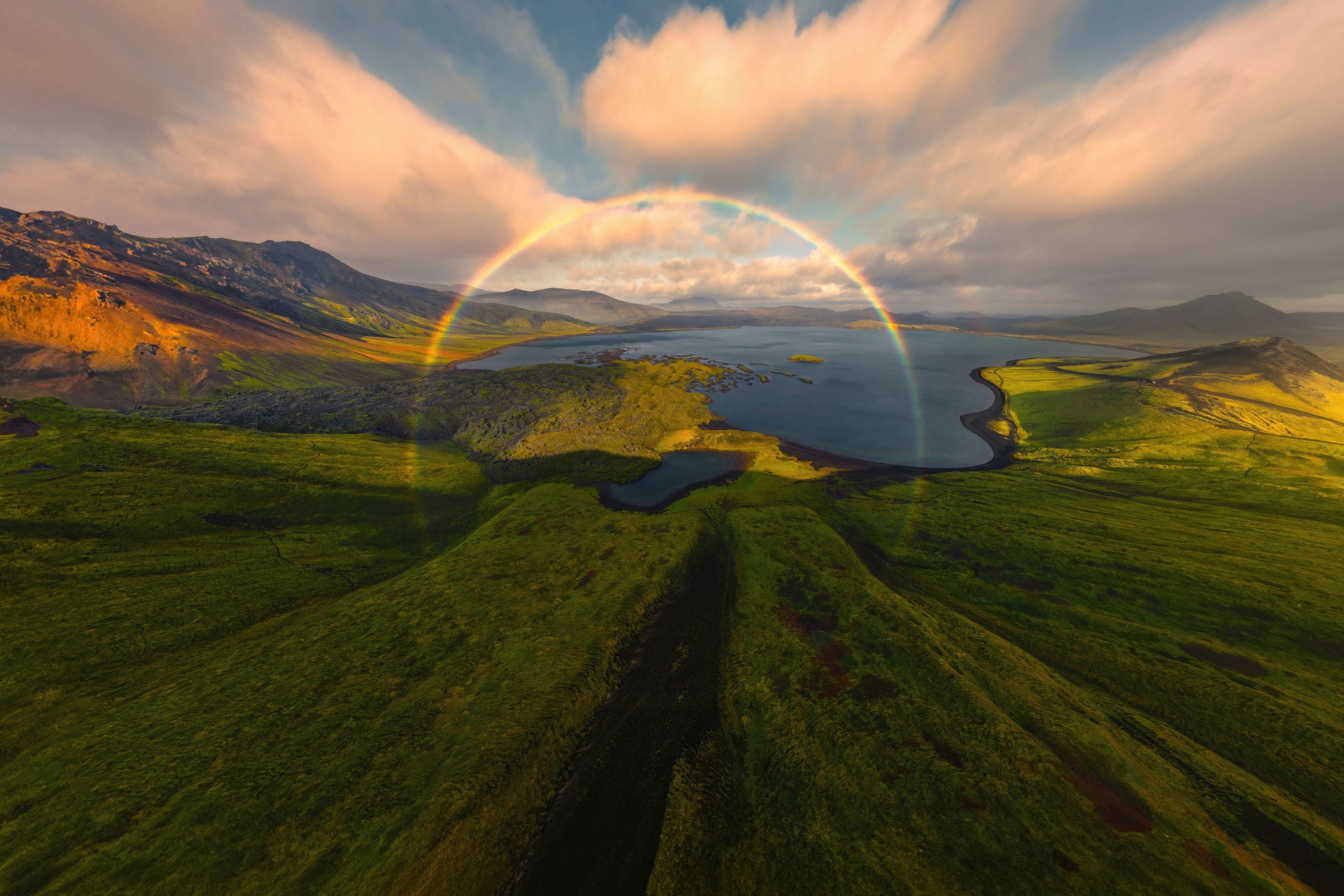 Ein leuchtender Regenbogen spannt sich über grünen Hügeln und einem ruhigen See in einer dramatischen Vulkanlandschaft.