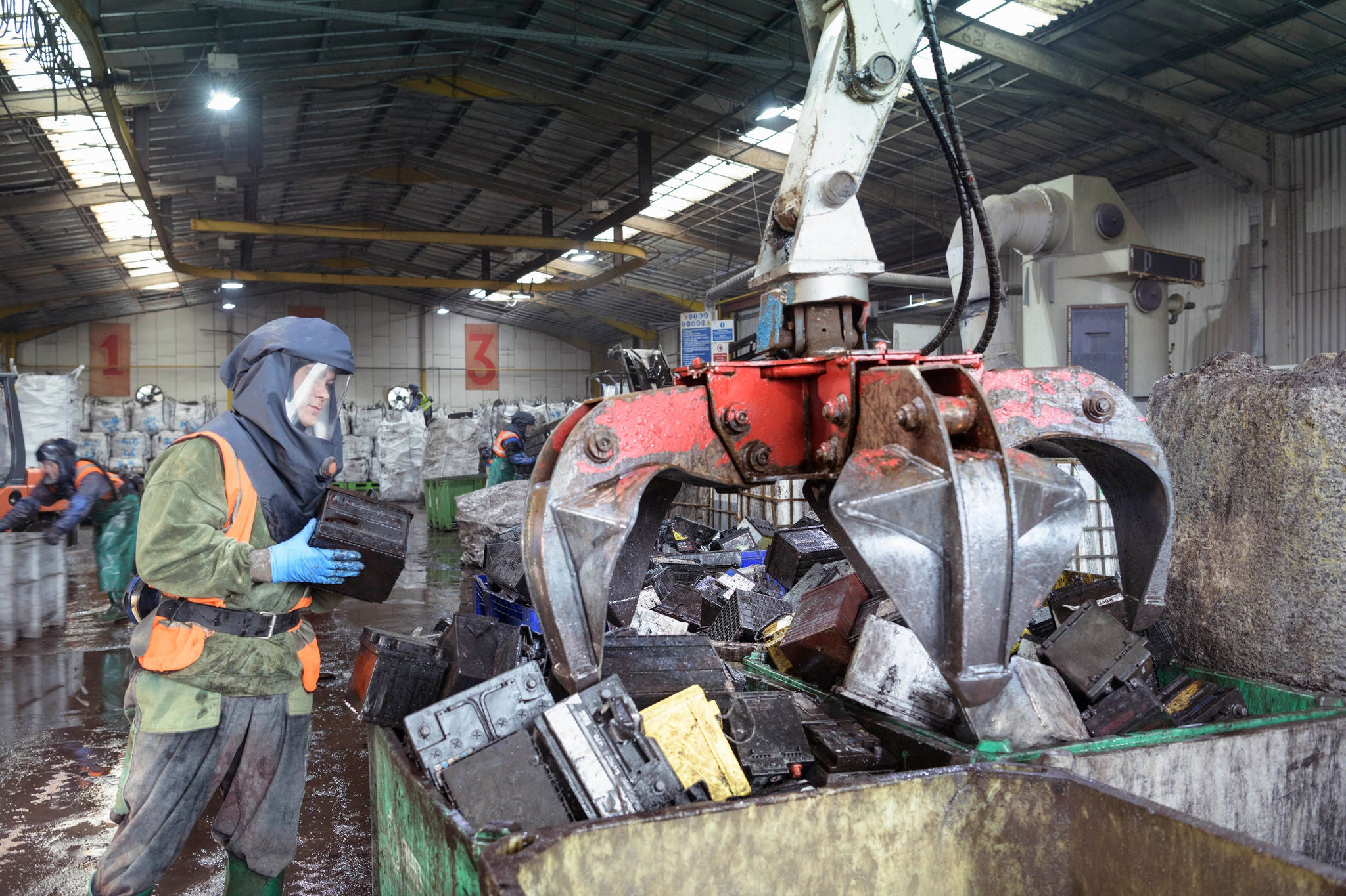 Worker in protective clothing loading vehicle batteries into grab bin in vehicle battery recycling plant