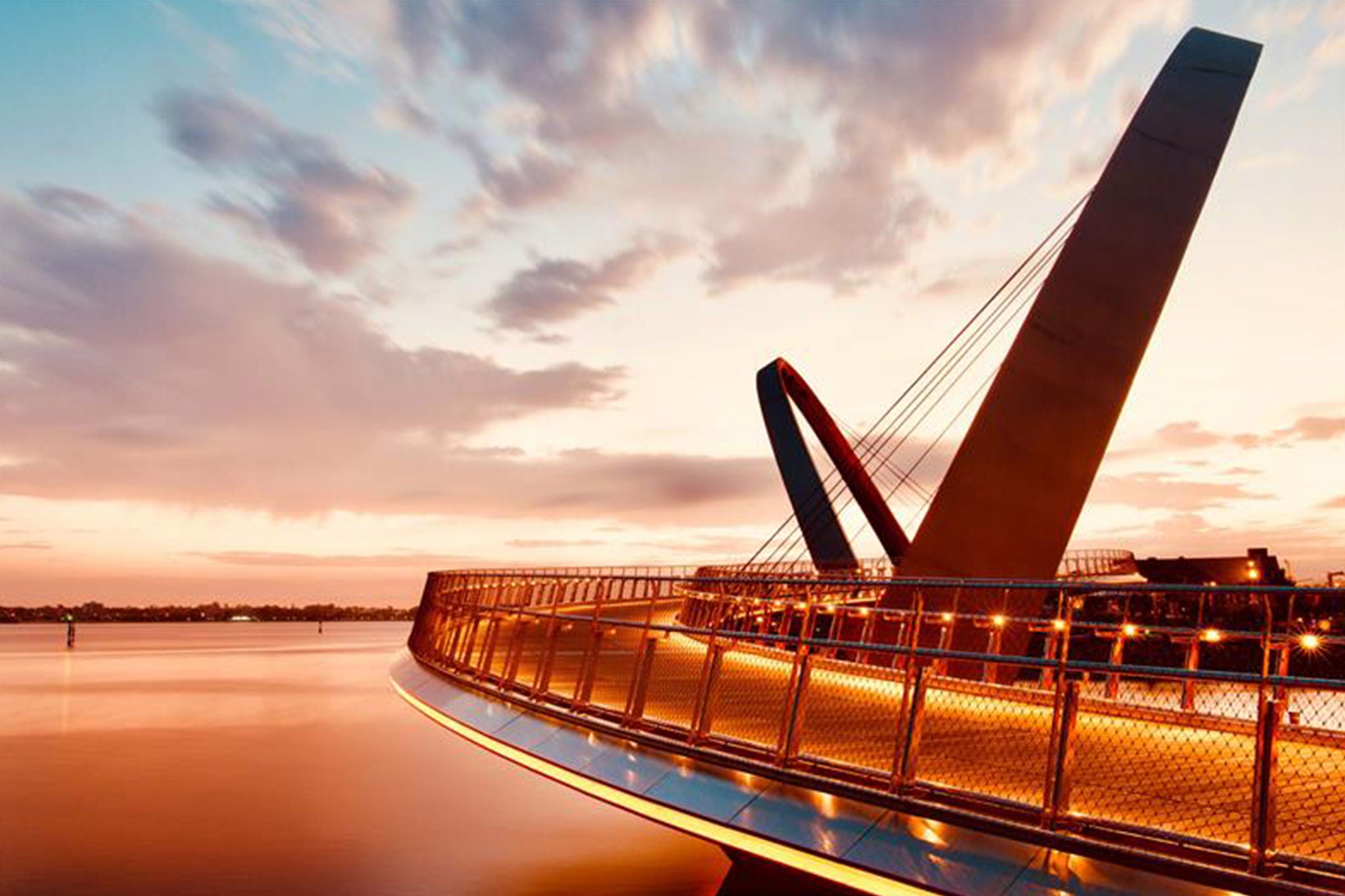 Curved pedestrian bridge with angled arches over water at sunset.