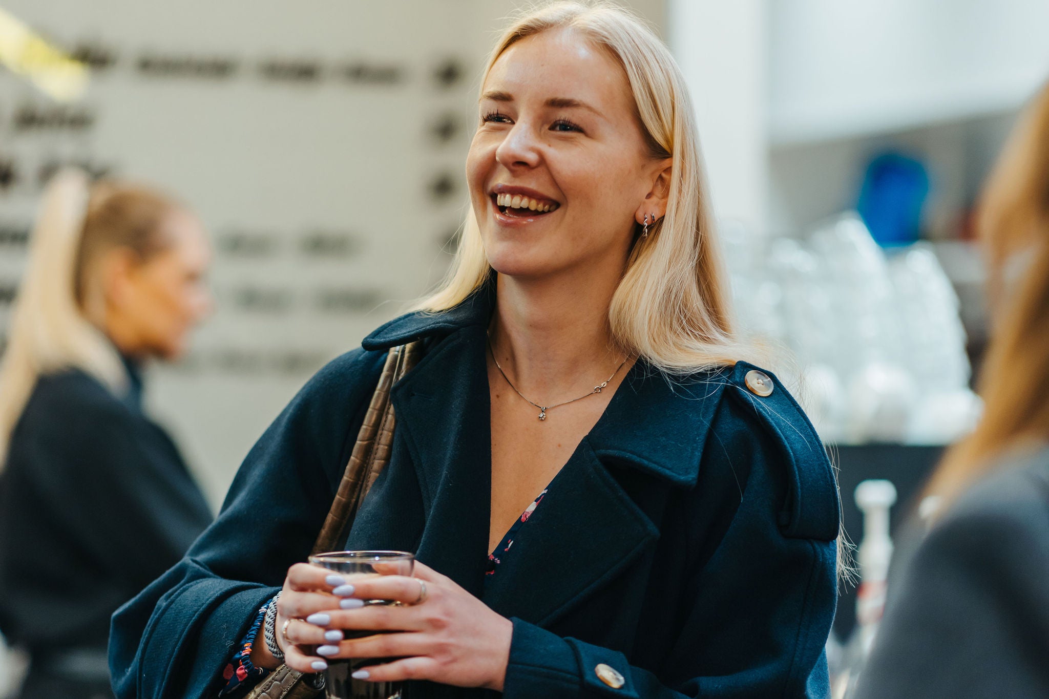 Woman holds a drink and talks in a social office setting, with other people in the background.